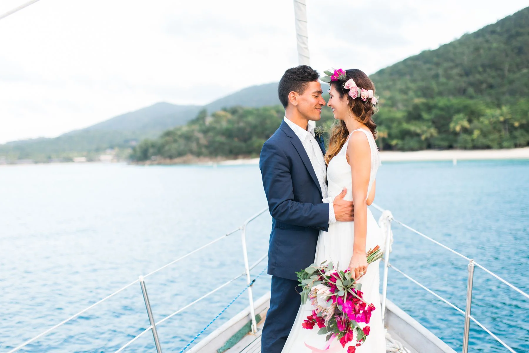 A bride dressed in white holds her brilliantly colored wedding bouquet and her groom aboard the sailboat Cimarron in St John, USVI.
