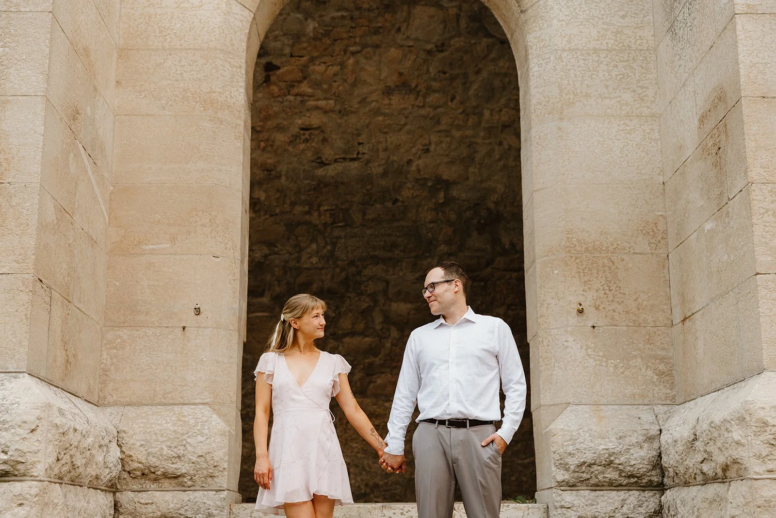  Couple holding hands under the stone arch at St. Boniface Cathedral in Winnipeg during an engagement session 