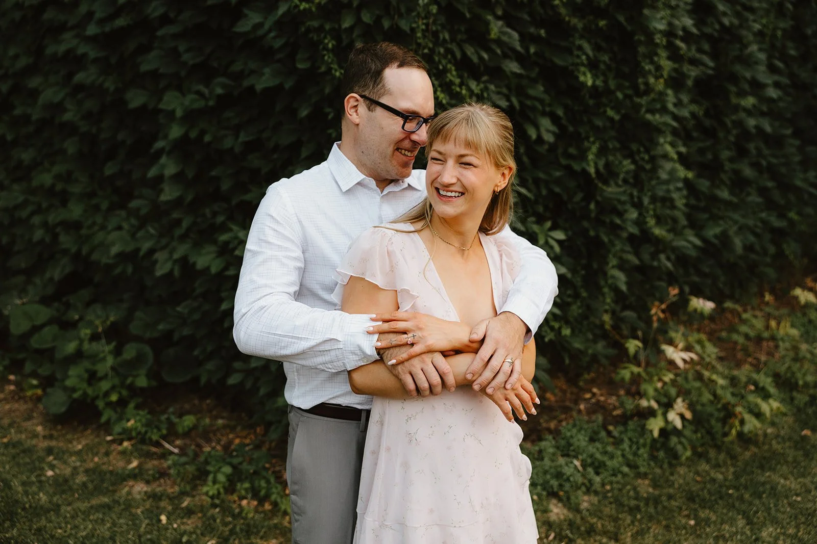  Couple laughing and embracing during an engagement session at St. Boniface Cathedral in Winnipeg with greenery backdrop 