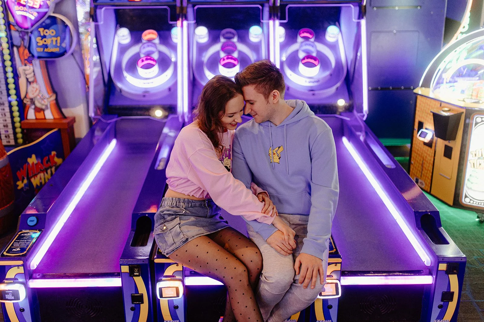  Couple sitting together at skeeball machines during a fun engagement session at The Rec Room in Winnipeg with neon arcade lights 