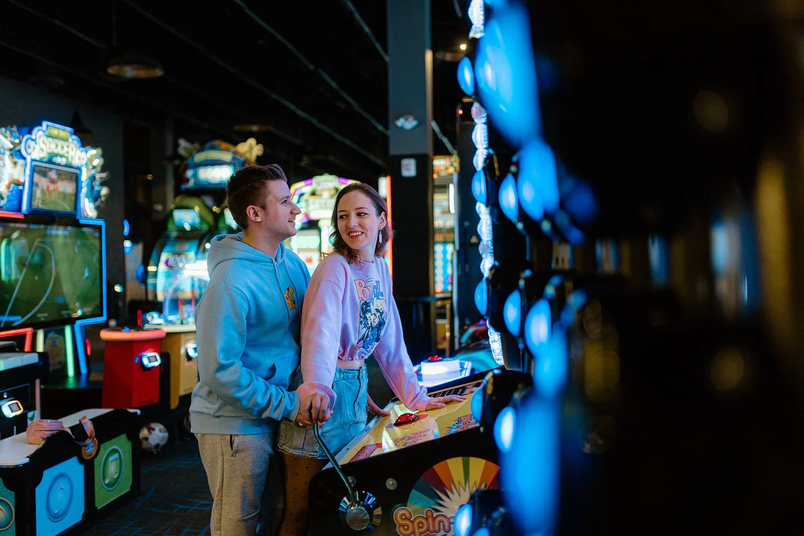  Couple smiling and playing arcade games during an engagement session at The Rec Room in Winnipeg with colorful neon lights 