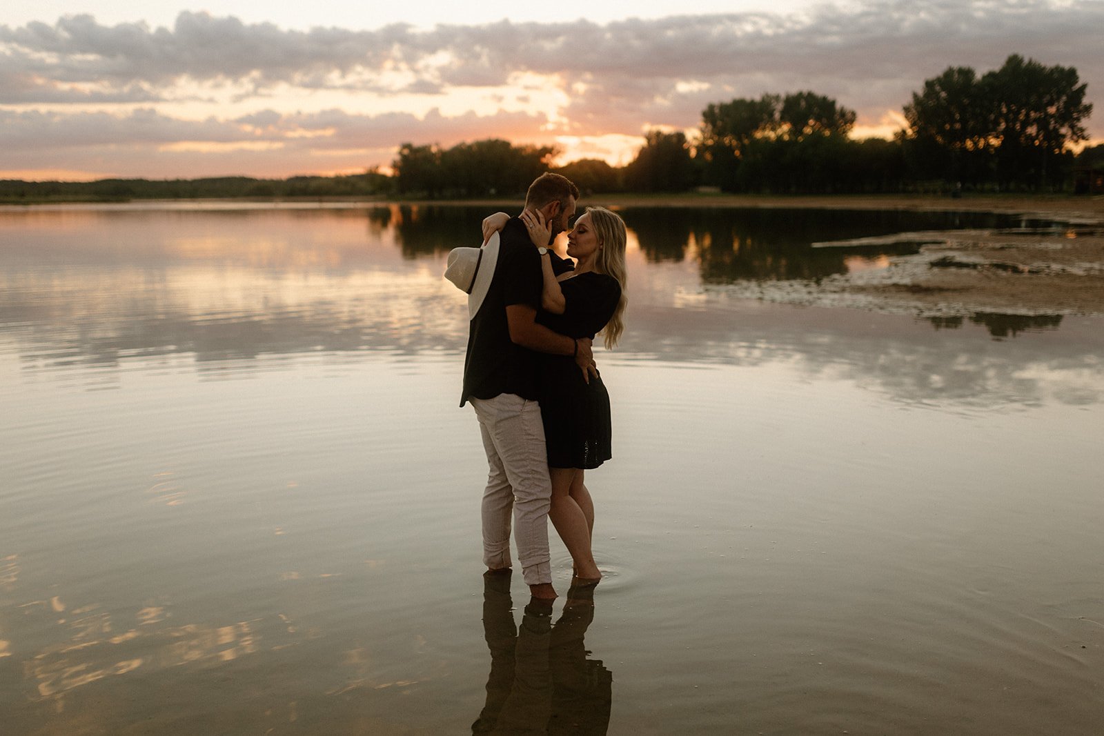  Couple embracing in shallow water at sunset in Birds Hill Park near Winnipeg with soft reflections on the lake   