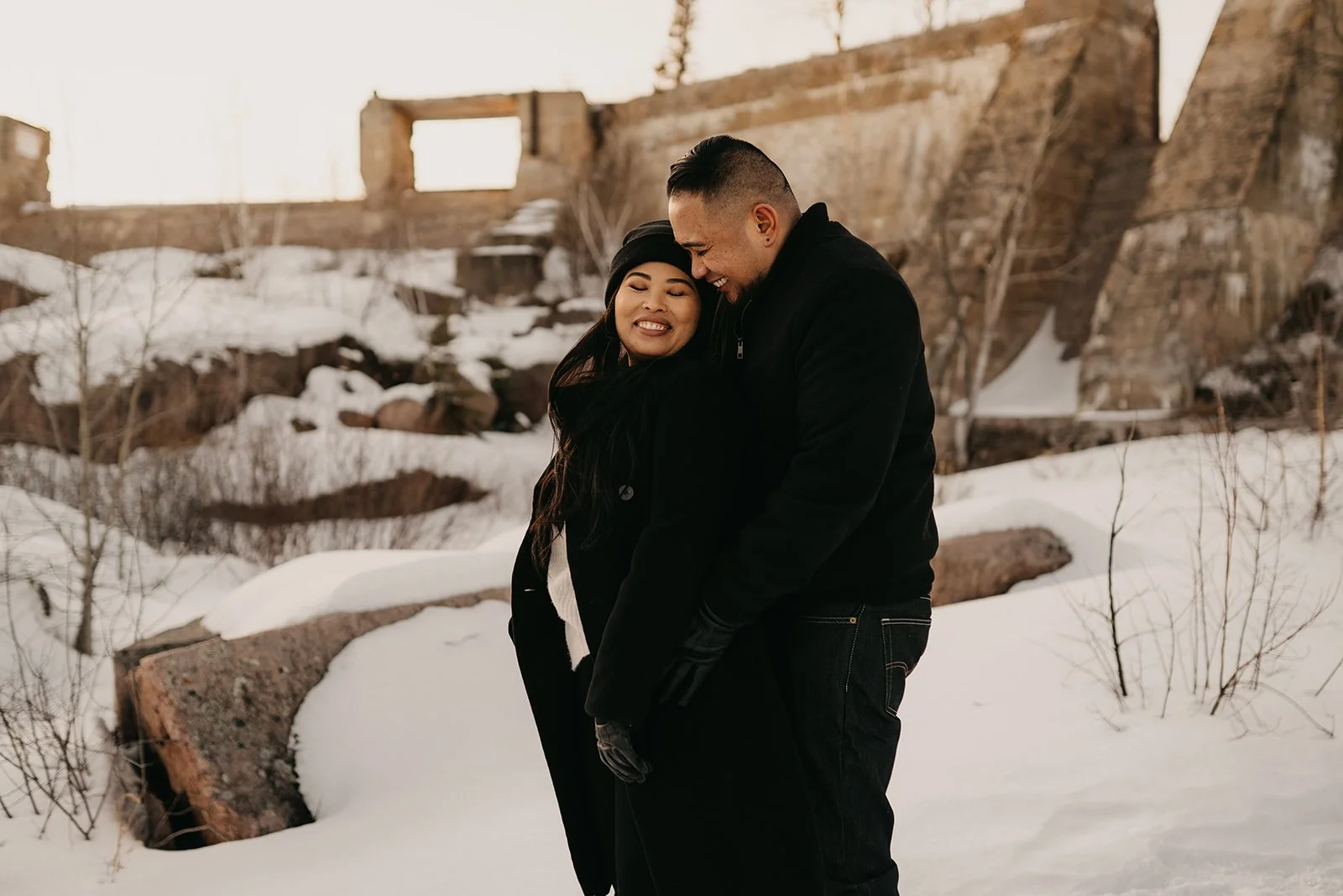  Couple embracing in the snow at Pinawa Dam in Manitoba during winter, with historic stone ruins in the background 