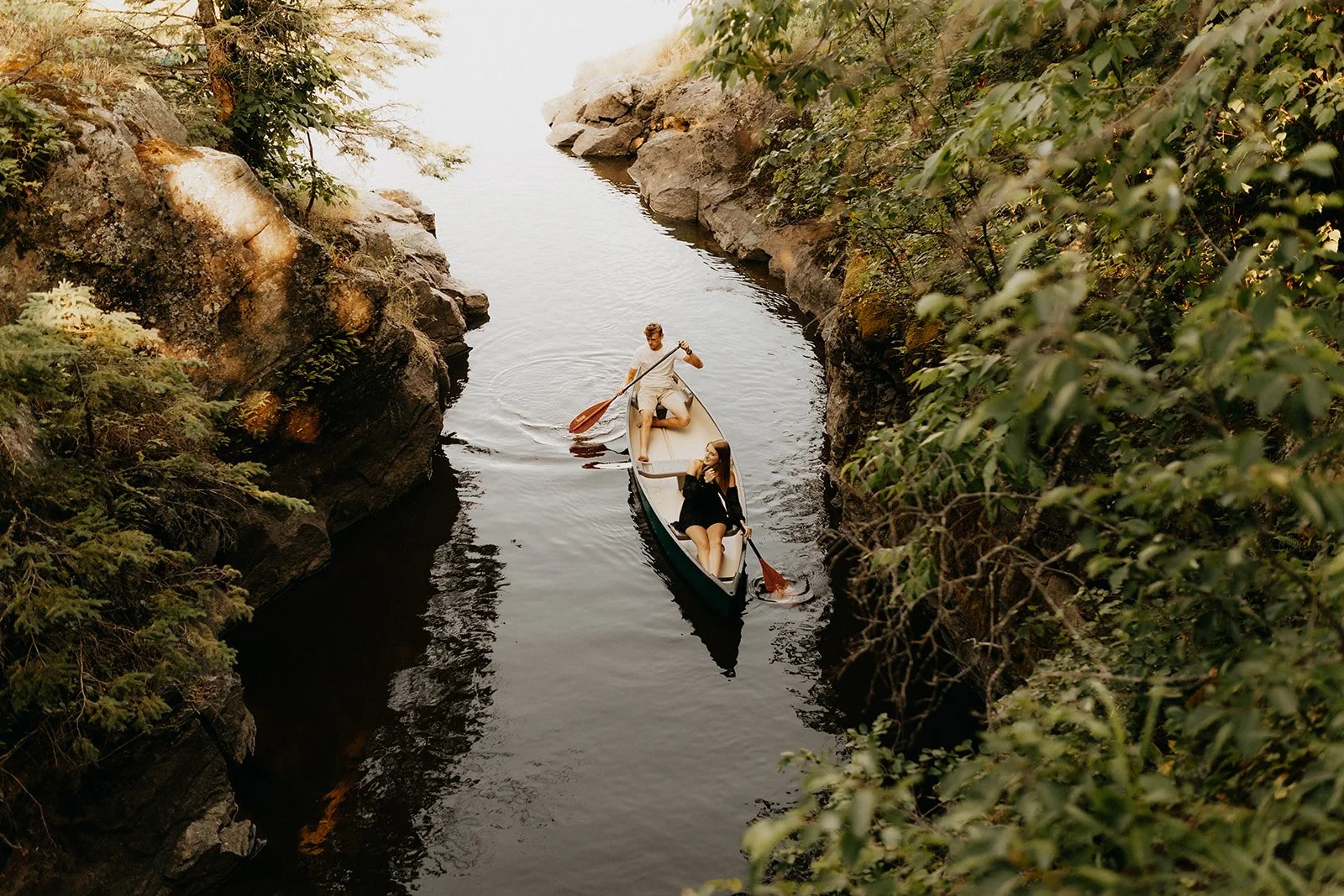 Couple paddling a canoe through a narrow rocky river in Whiteshell Provincial Park, surrounded by trees and natural landscape 