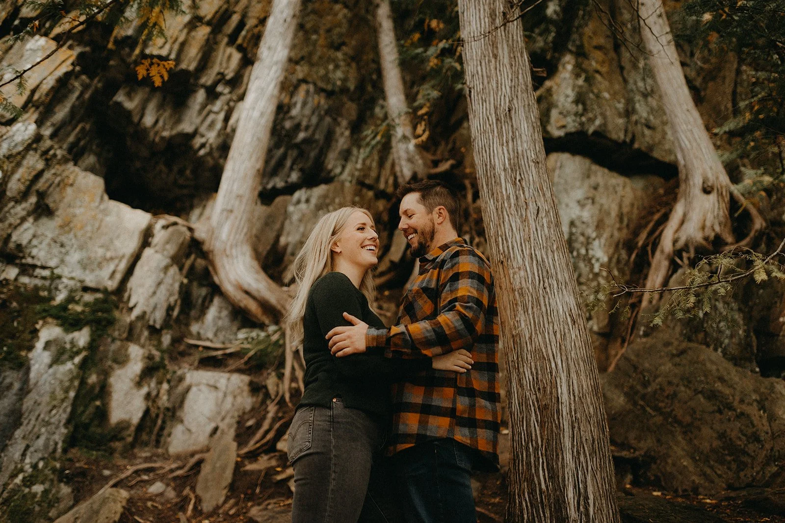  Couple embracing and laughing in front of rugged rock cliffs and trees at Hunt Lake in Whiteshell Provincial Park Manitoba 