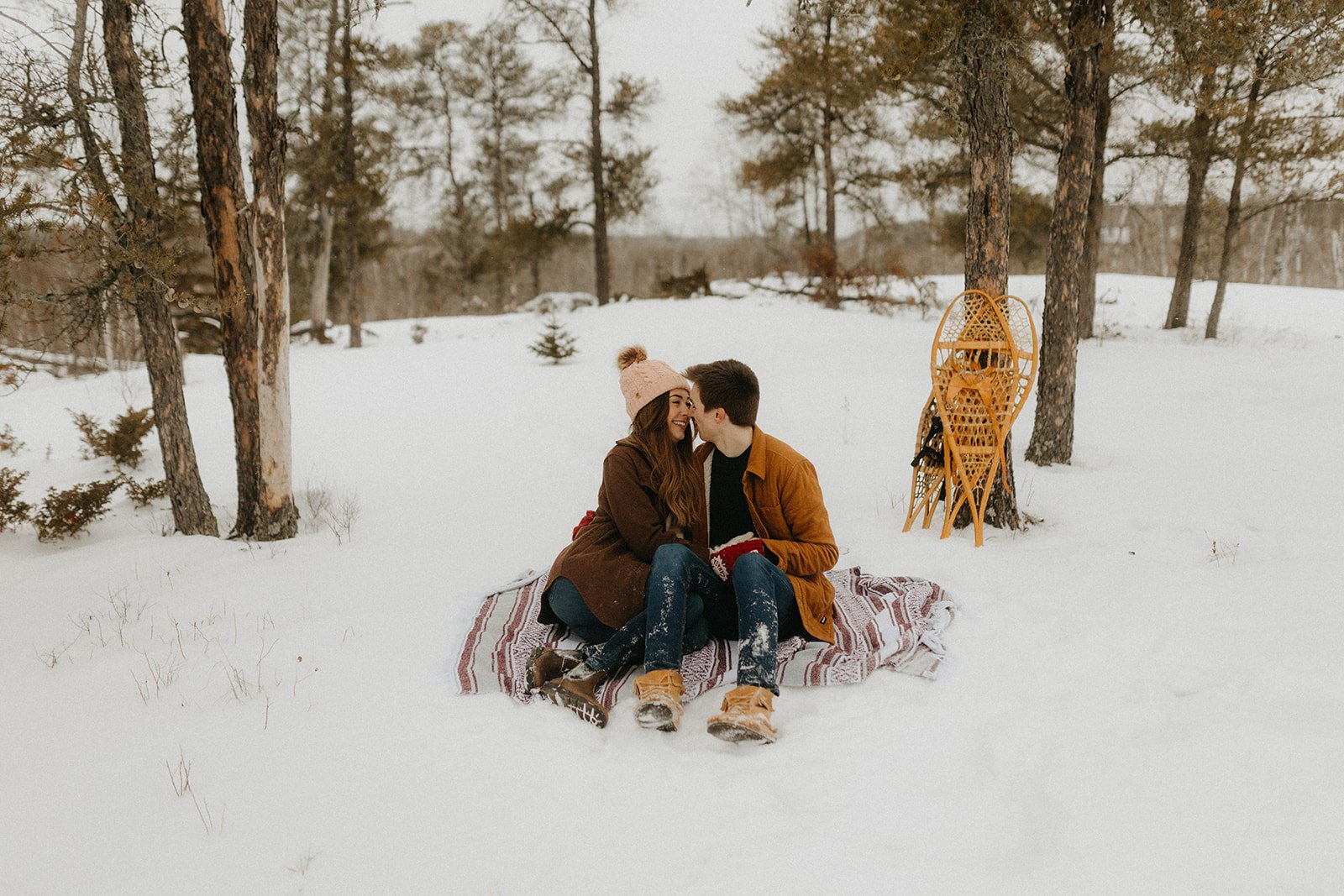  Couple sitting on a blanket in the snow, laughing together among pine trees during a winter engagement session in Whiteshell Manitoba 
