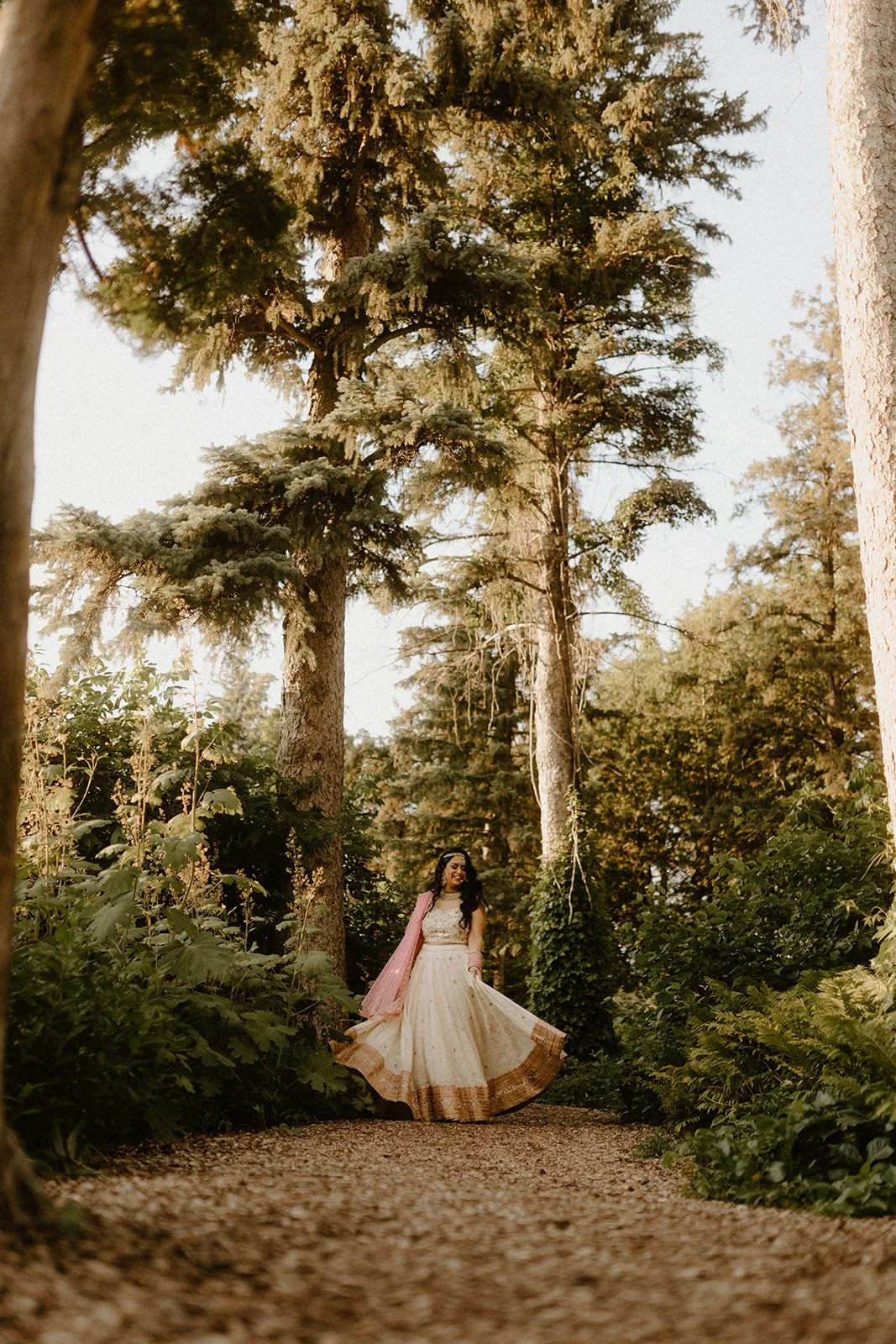  Woman twirling in a traditional South Asian outfit on a garden path in the English Garden at Assiniboine Park during a pre wedding photoshoot in Winnipeg, surrounded by tall trees and lush greenery 