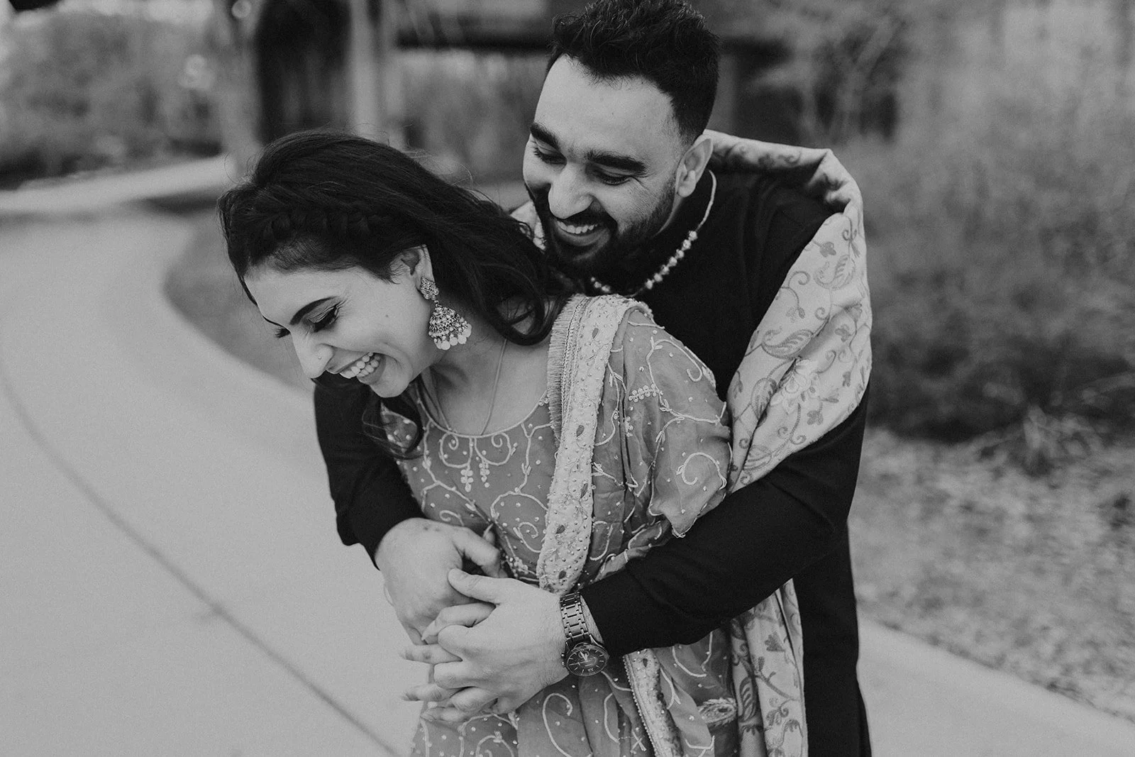  Couple laughing together beneath the CN Rail bridge at The Forks during a pre wedding photoshoot in Winnipeg, wearing traditional South Asian outfits 