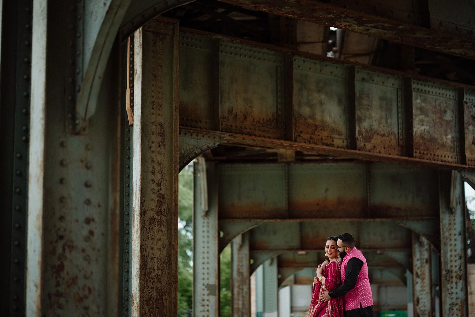  Couple embracing beneath the CN Rail bridge at The Forks during a pre wedding photoshoot in Winnipeg, wearing traditional South Asian outfits among rusted steel beams 