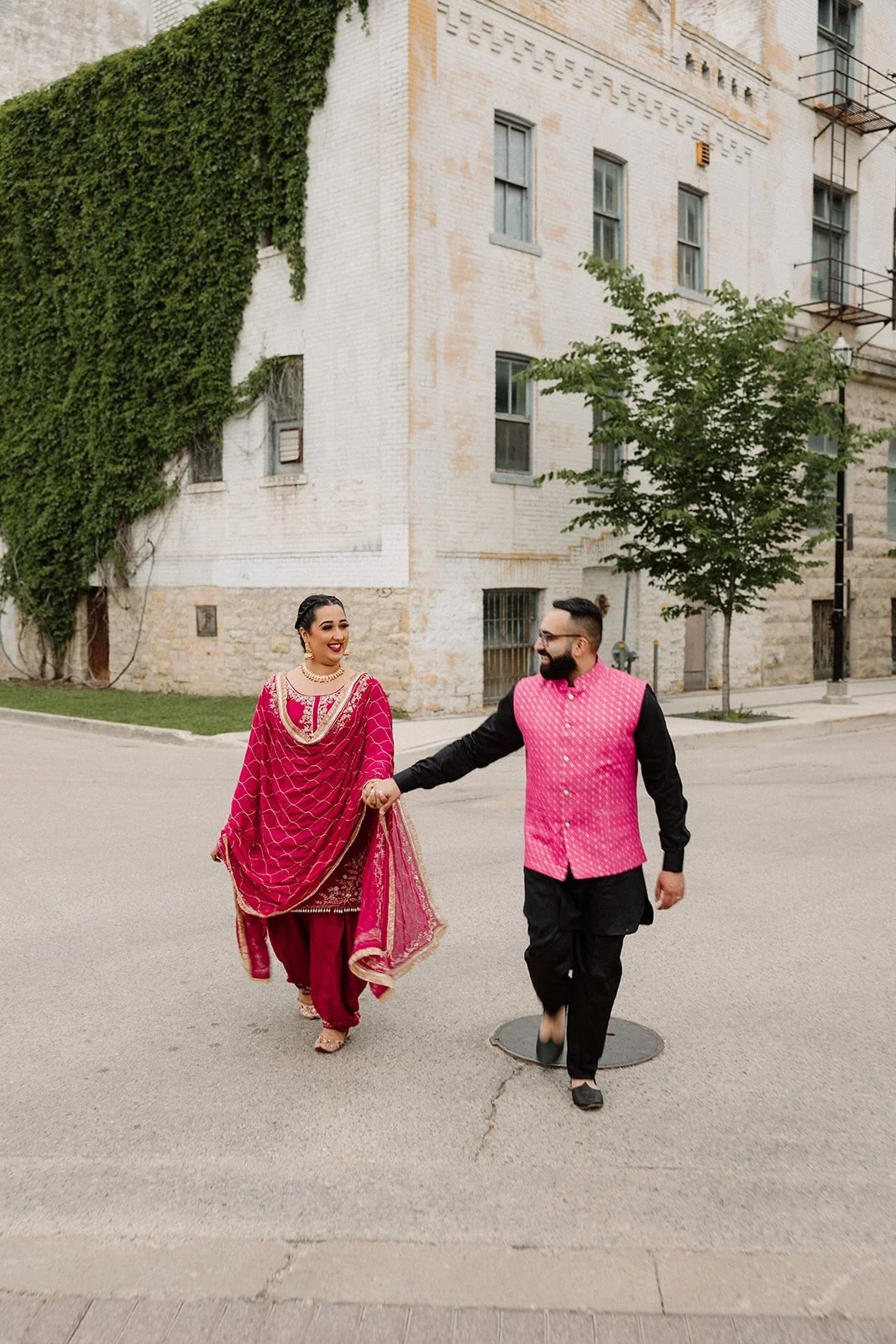  Couple walking together during a pre wedding photoshoot in Winnipeg’s Exchange District, wearing traditional South Asian outfits and holding hands 