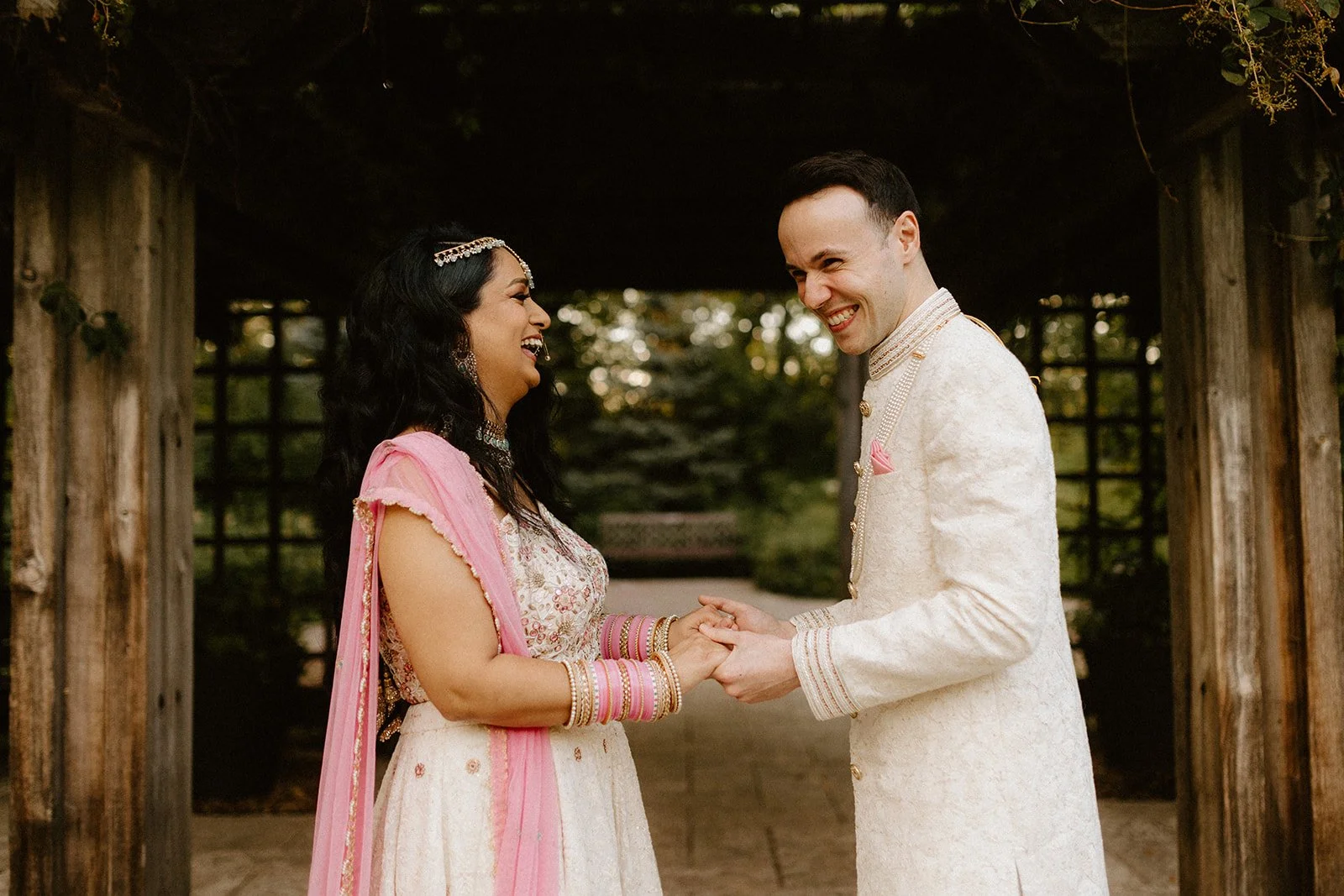  Couple laughing and holding hands during a pre wedding photoshoot in Winnipeg, wearing traditional South Asian outfits in a garden setting 