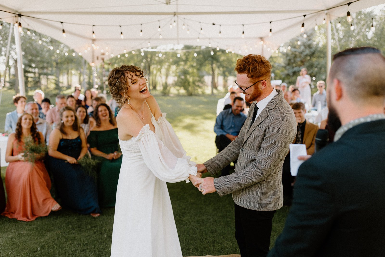  Bride laughing during a backyard tent wedding ceremony in southern Manitoba, surrounded by guests seated on the grass 