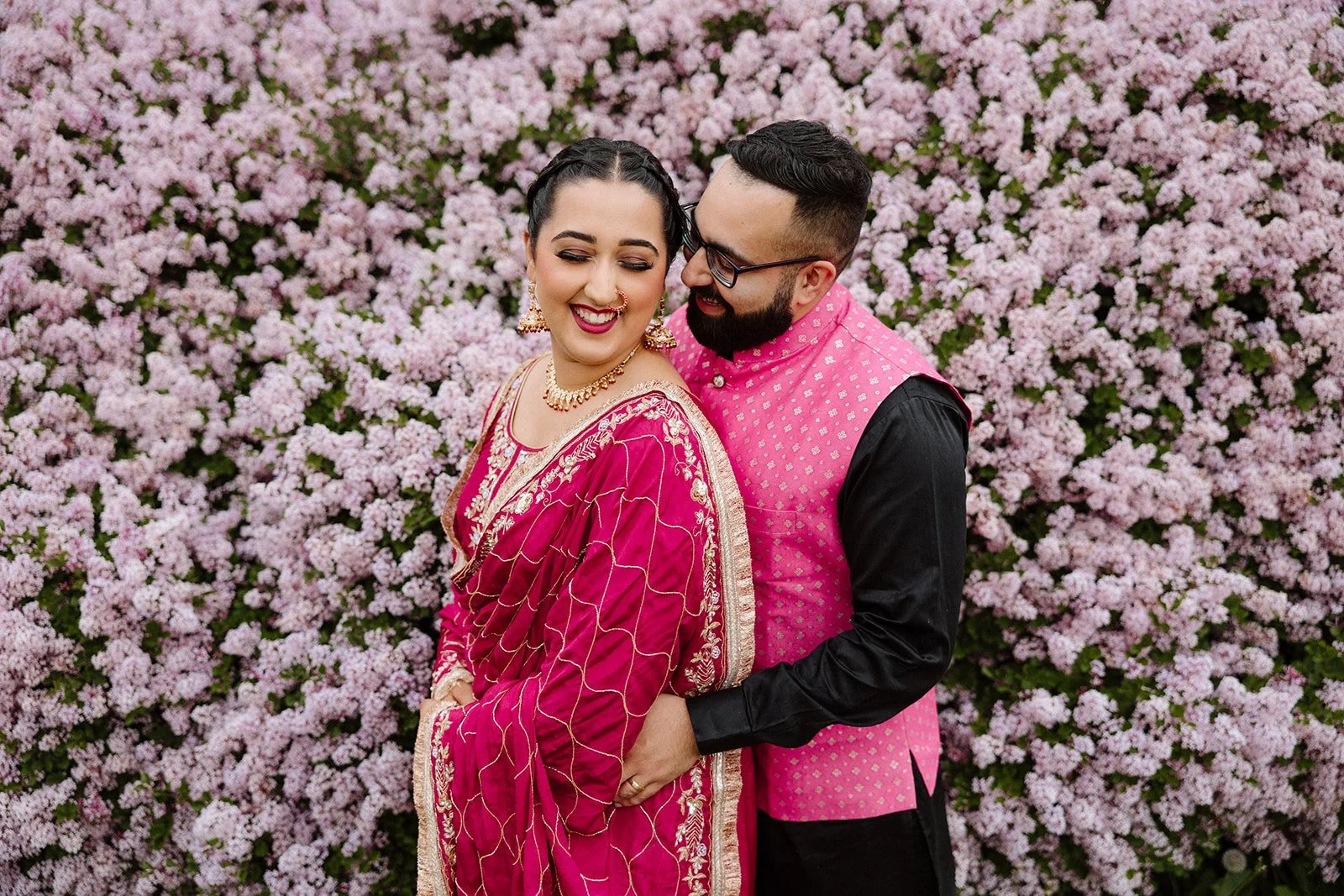 Couple laughing together during a pre wedding photoshoot in Winnipeg, wearing traditional South Asian outfits in front of a wall of pink lilac blooms
