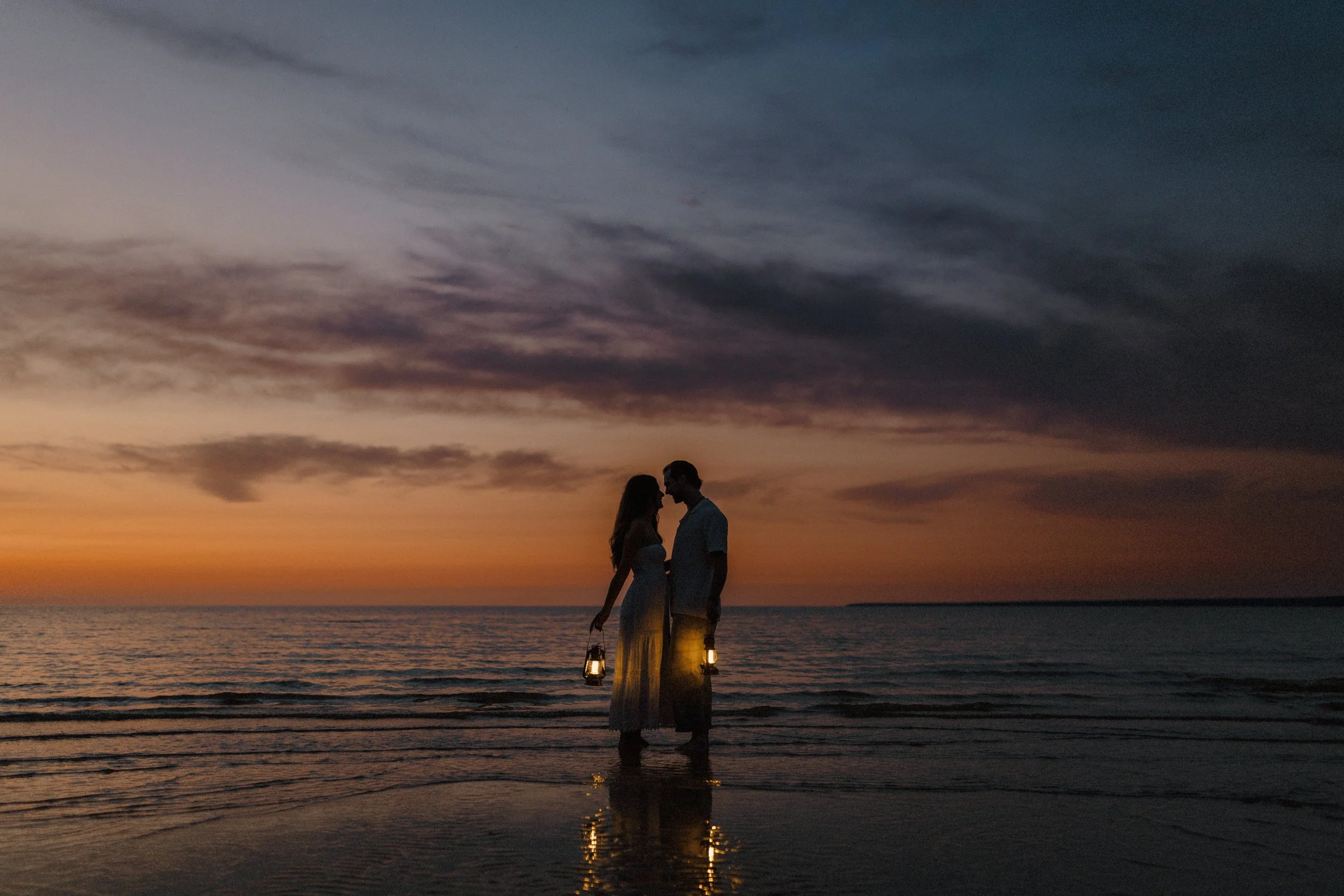 Couple standing in shallow water at sunset during a Grand Beach engagement session, holding lanterns at twilight.