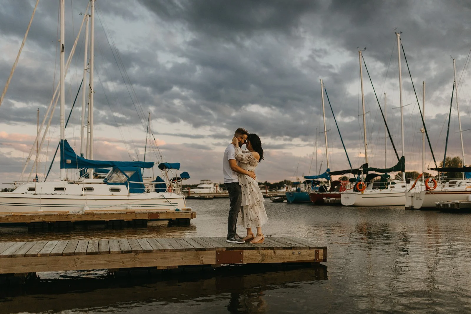 Couple kissing on a dock at Gimli Harbour on Lake Winnipeg with sailboats and dramatic evening sky