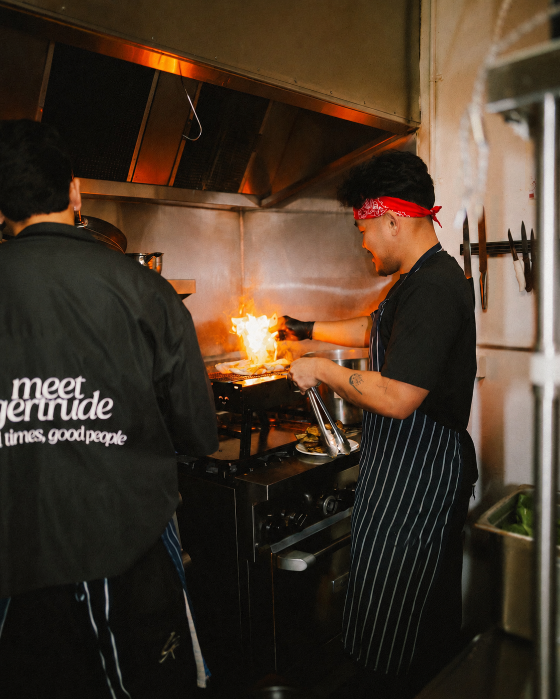 Person cooking on a stove with a flame, wearing a red bandana and striped apron, in a commercial kitchen.