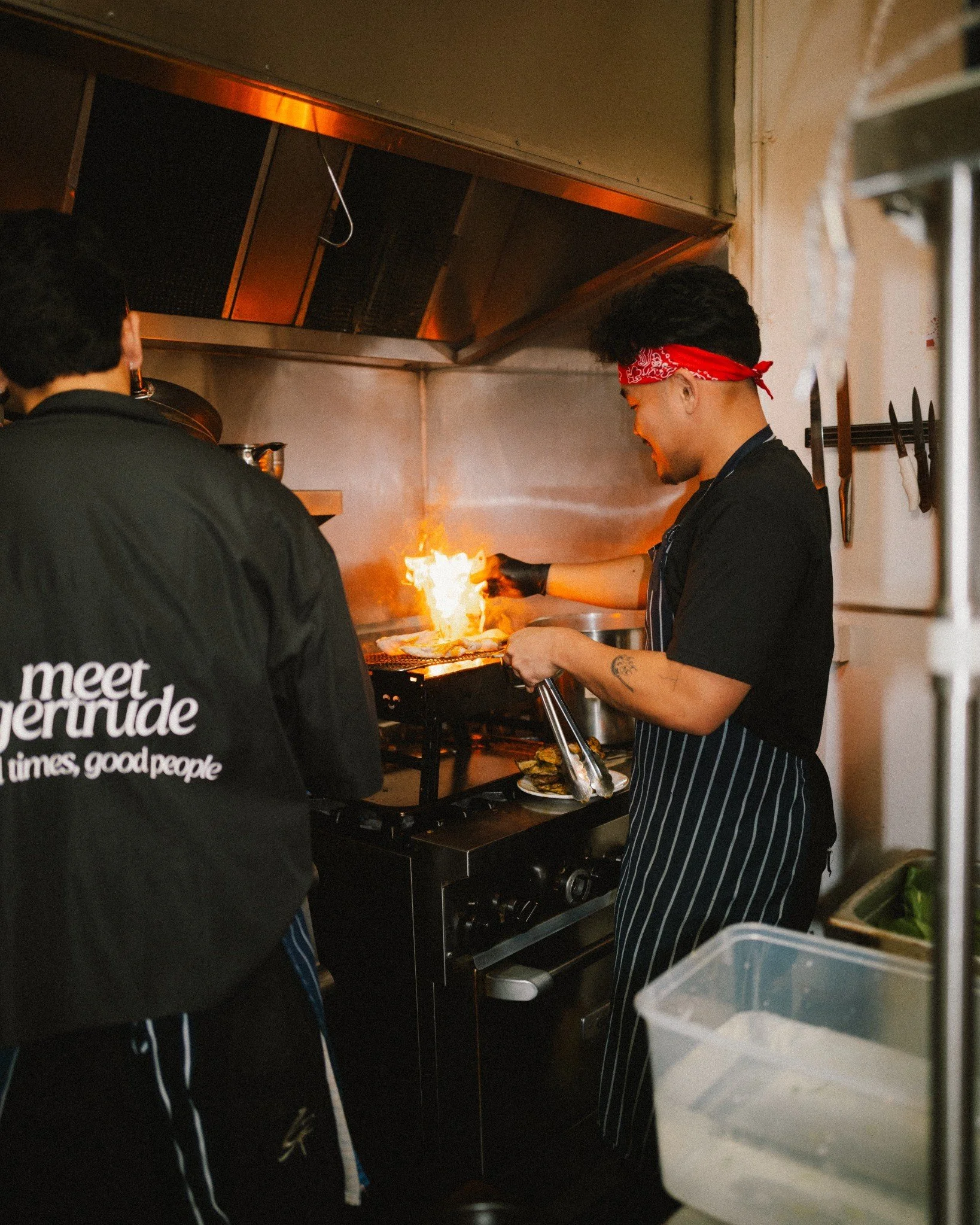 Two chefs working in a restaurant kitchen, one wearing a red bandana, black shirt, and striped apron, cooking over an open flame, with various kitchen tools and equipment visible around.