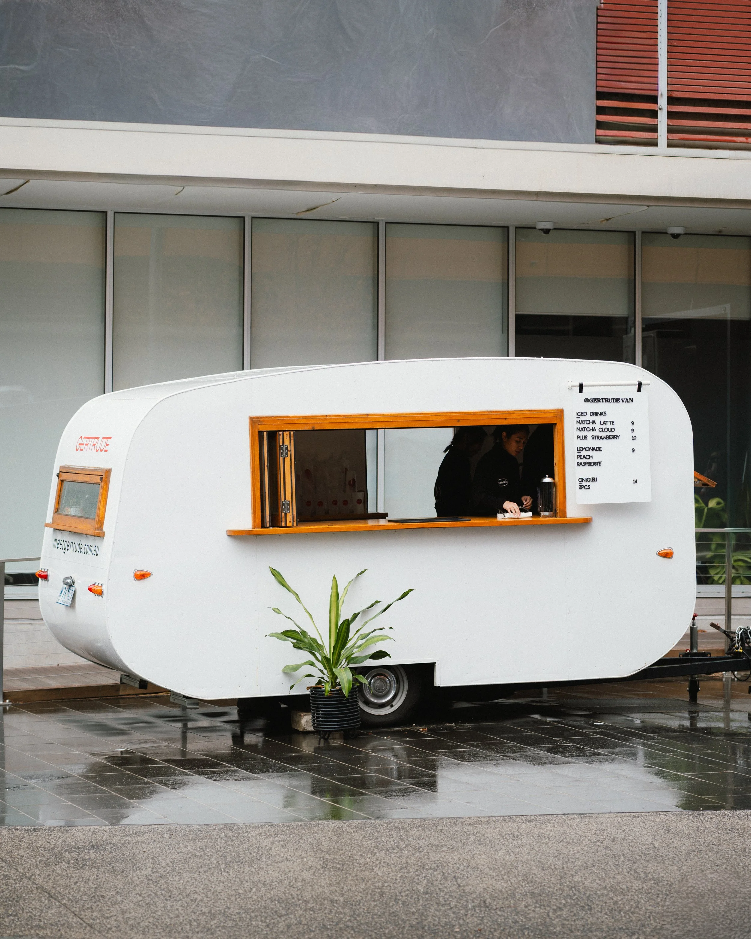 A white food truck with a wooden window serving counter, parked on wet tiled ground outside a modern building. There is a green potted plant in front of the truck. The menu on the side of the truck lists drinks and food items.