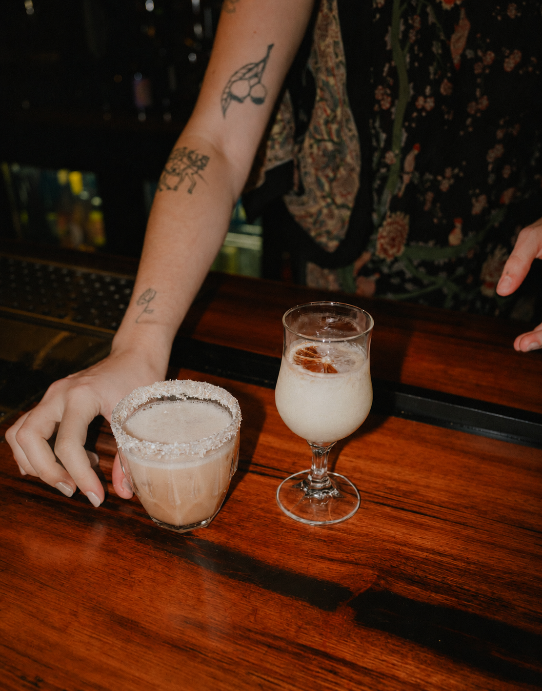 Two cocktails on a wooden bar: a frosted glass of frozen margarita with salt rim and a tall glass of creamy tropical drink with ice and a cherry garnish. A woman's hand and arm with tattoos are reaching for the margarita.