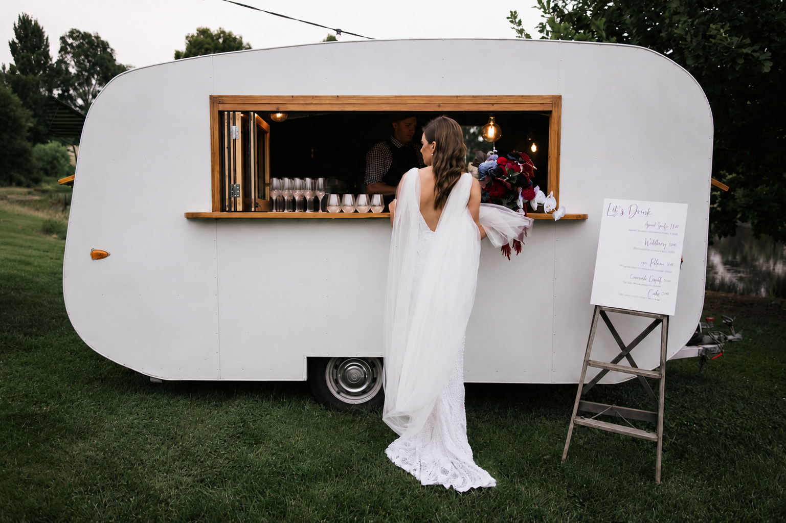 Bride in a white wedding dress and veil ordering drinks at a mobile bar trailer during outdoor event.