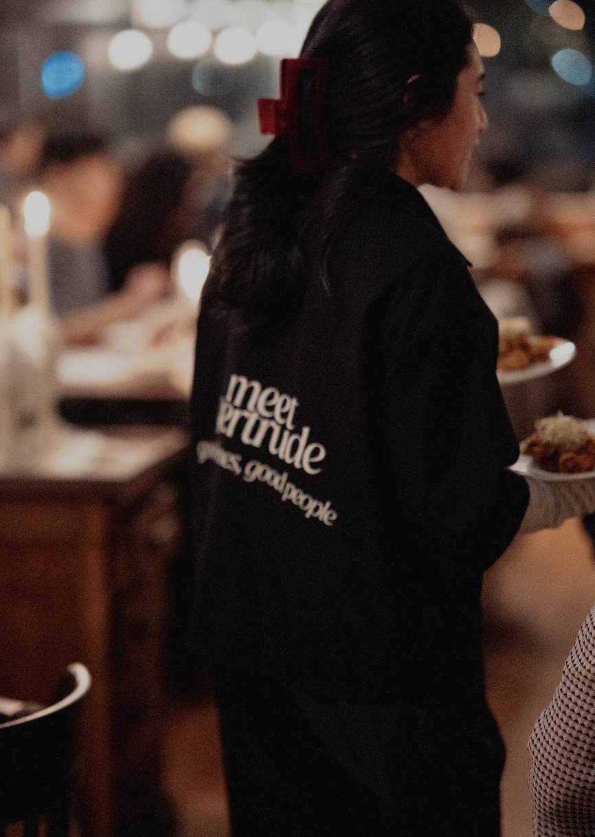 A woman wearing a black shirt with white text that says "meet new people, make good people" stands at a dining event, holding a plate of food, with a busy restaurant or event in the background.