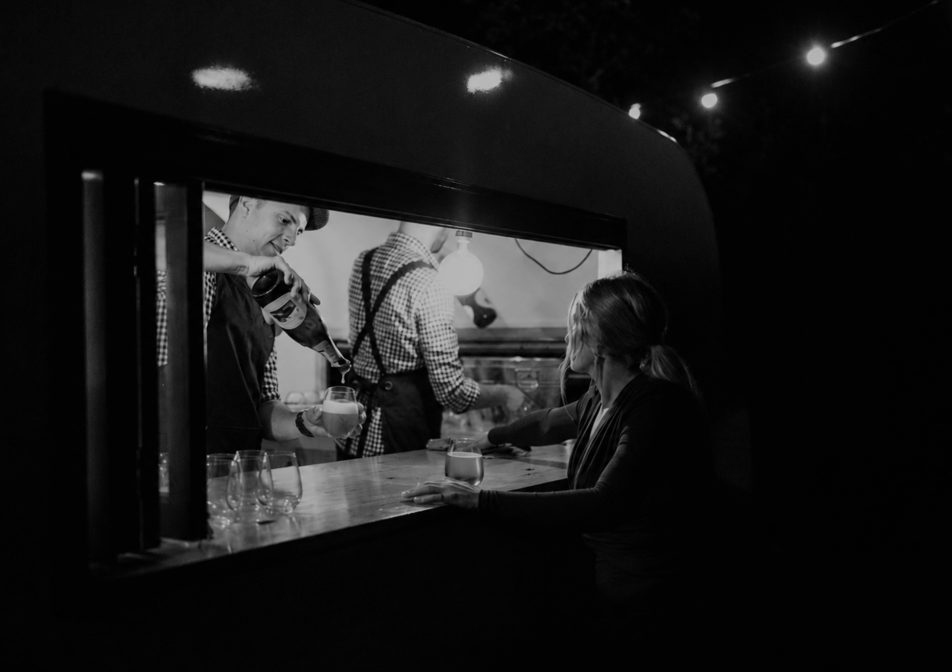 Black and white photo of a man pouring drinks at a bar during nighttime, viewed through a window. The scene features people engaging in conversation.