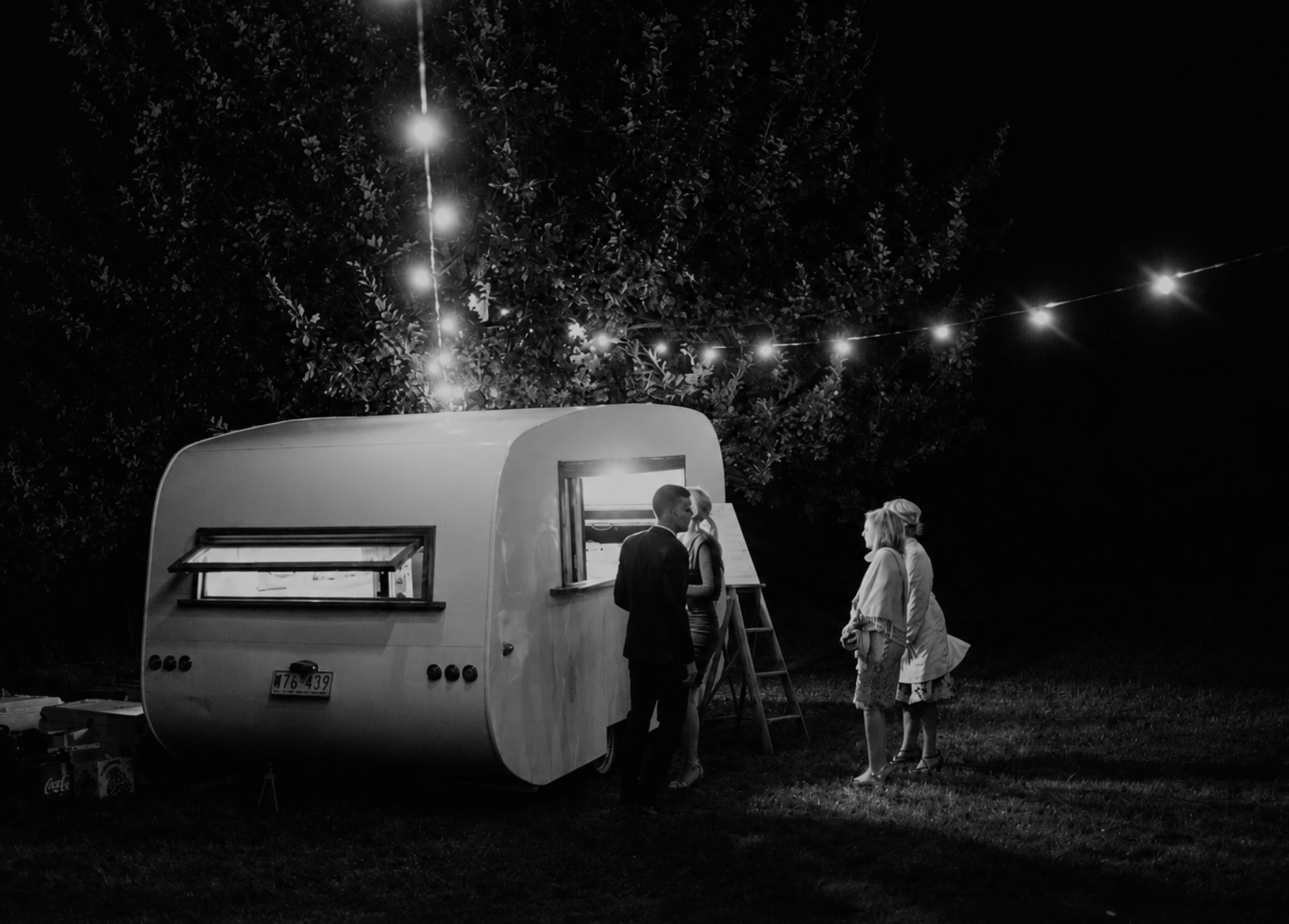 A black-and-white photo shows three people standing outside a vintage food truck at night. String lights hang above them, illuminating the scene, with trees in the background.