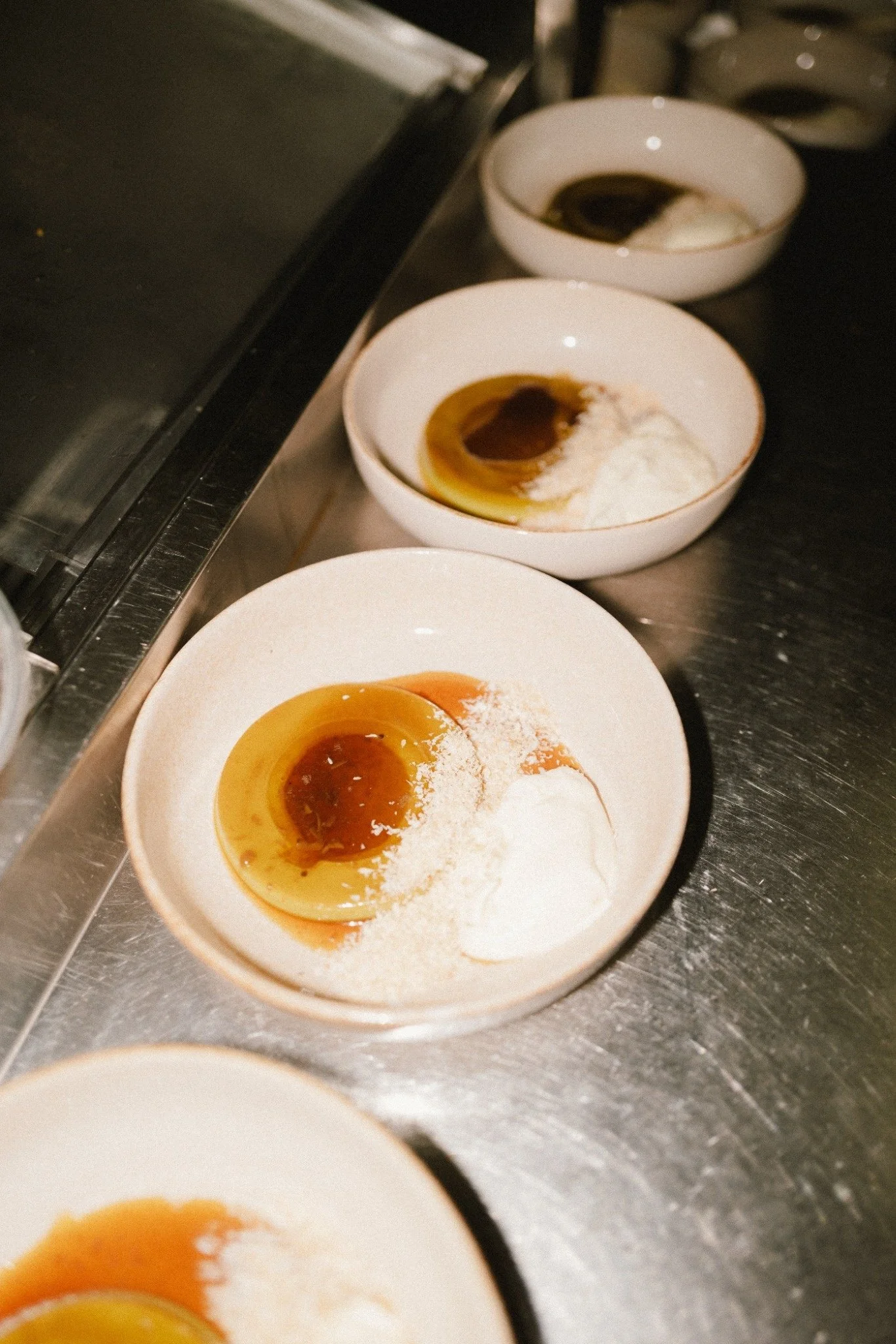 Three white bowls of caramel flan with whipped cream on top, placed on a metal counter in a kitchen.