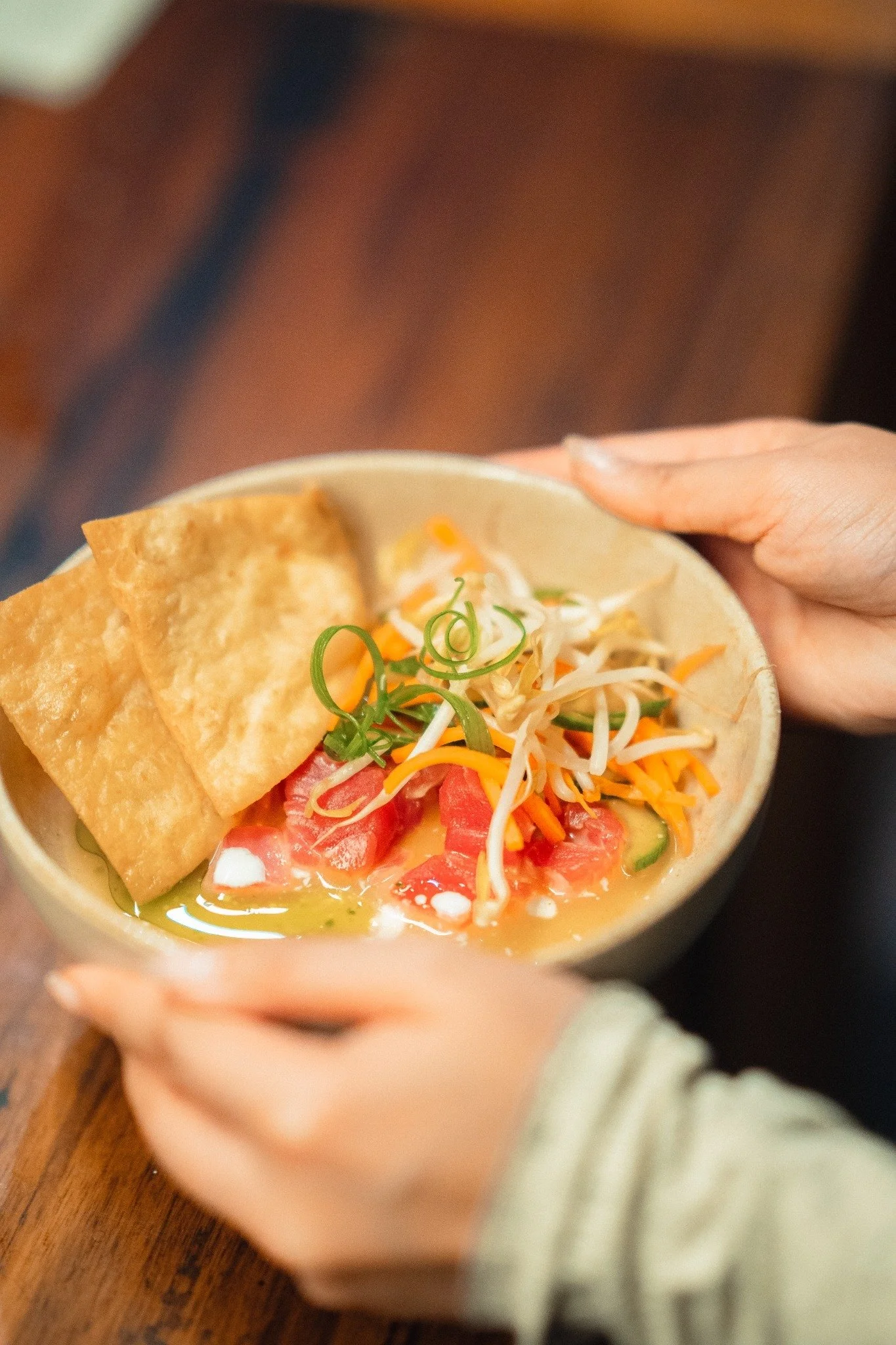 A person holding a bowl of ramen with fried tofu, shredded vegetables, green onions, and a soft-boiled egg, with a wooden table surface underneath.
