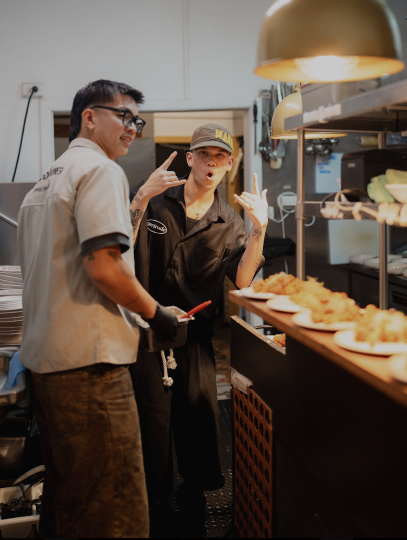 Two chefs in a restaurant kitchen making playful poses in front of a tray of fried food.