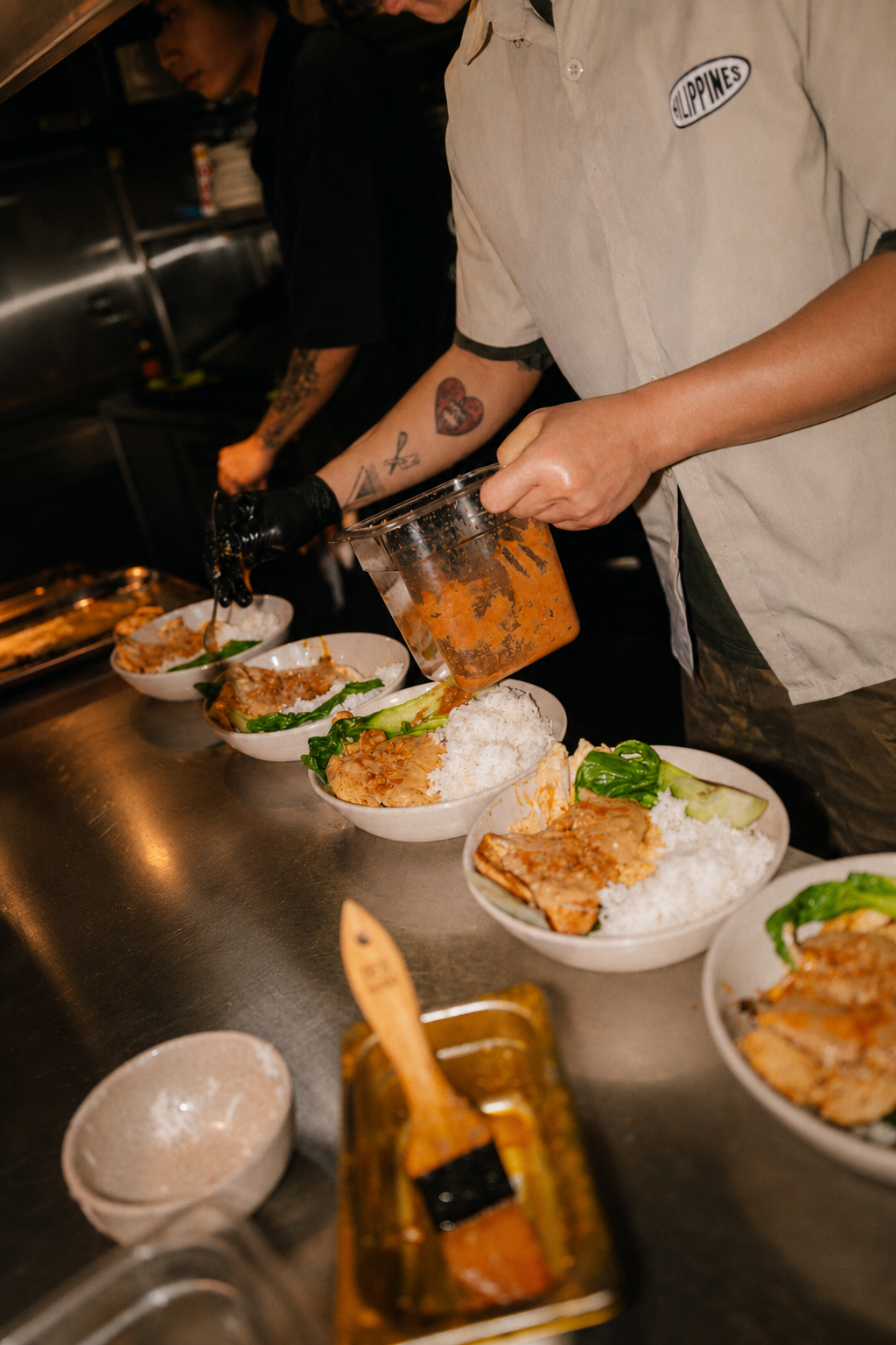 A chef pouring sauce over bowls of rice and vegetables in a busy kitchen.