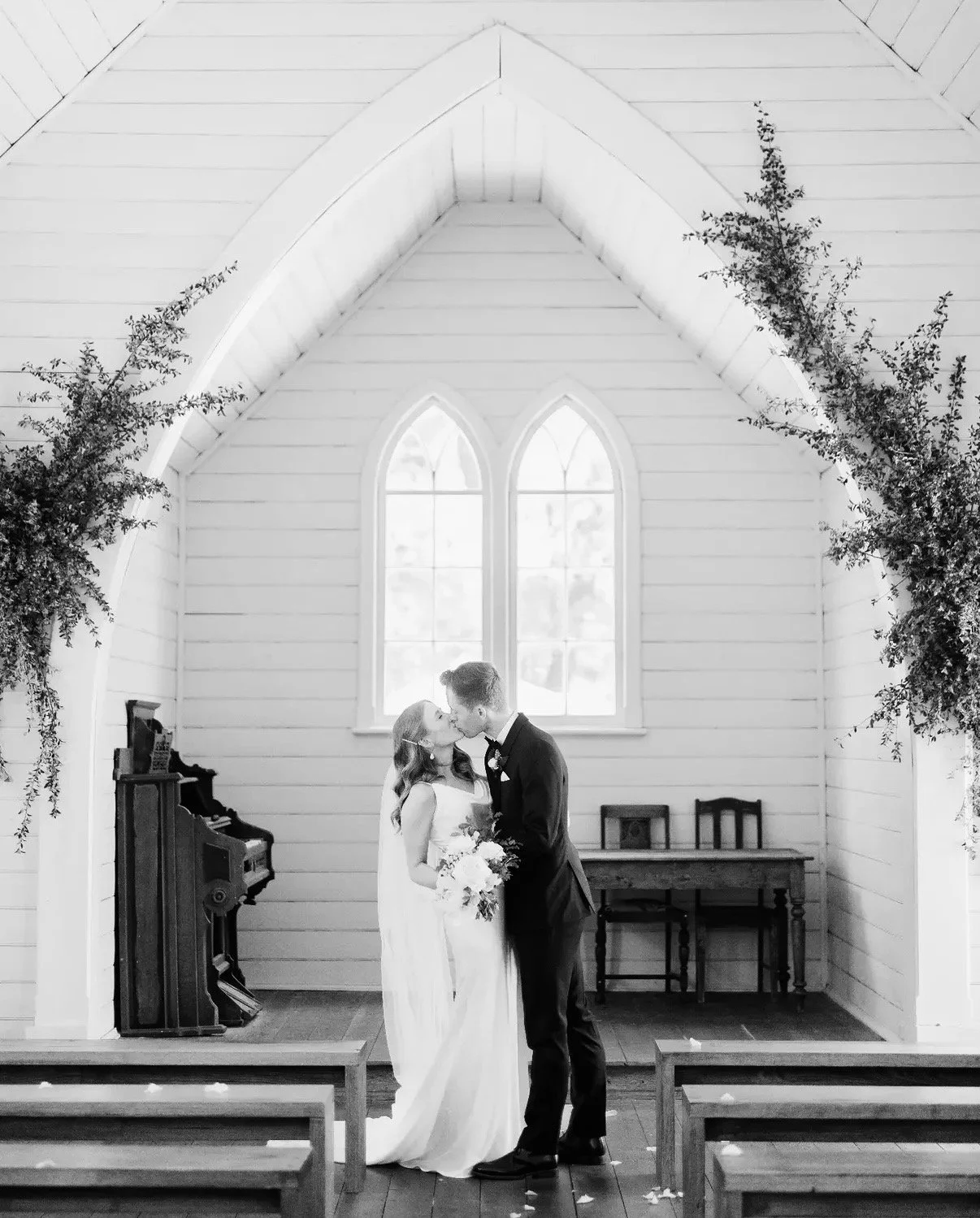 A bride and groom kissing inside a small wooden church with white walls and large arched windows, decorated with greenery on either side.