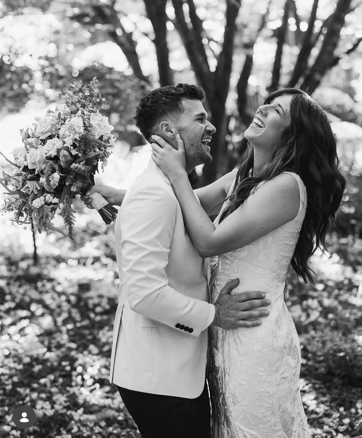 A black and white photo of a joyful couple at their wedding, outdoors, with the woman holding a bouquet of flowers and both smiling and embracing each other.