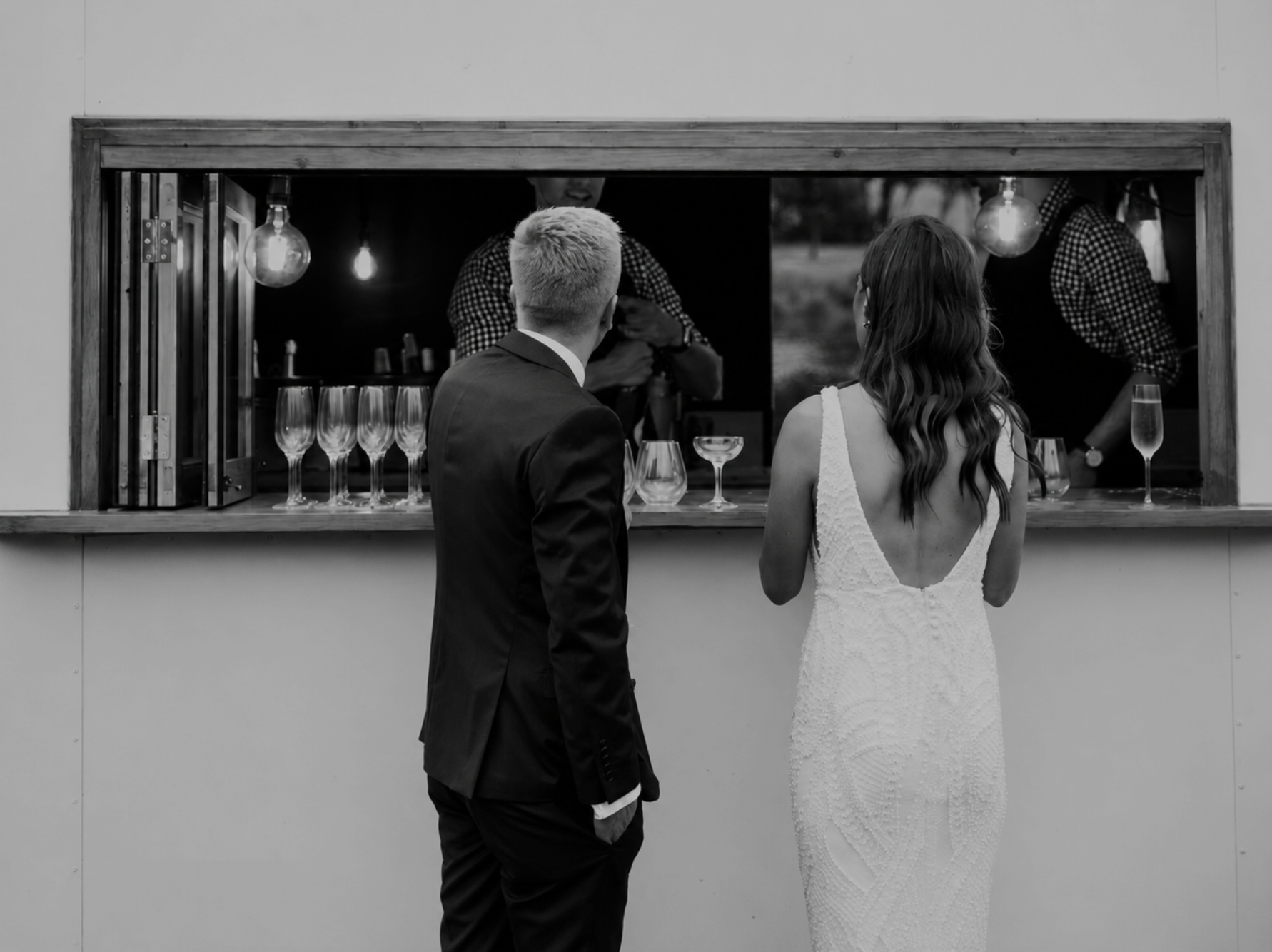 A black-and-white photo of a bride and groom standing in front of a food truck window, with the groom in a suit and the bride in a white wedding dress, as two people inside the truck prepare drinks.
