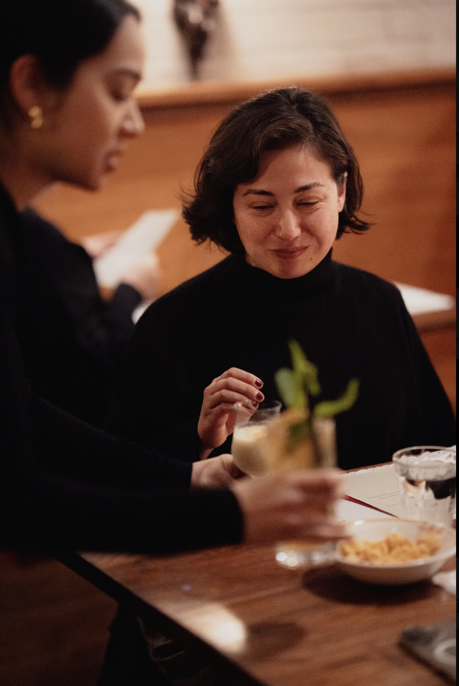 Two women sitting at a dinner table, one handing a drink to the other, with food and water glasses on the table in a warm-toned restaurant setting.