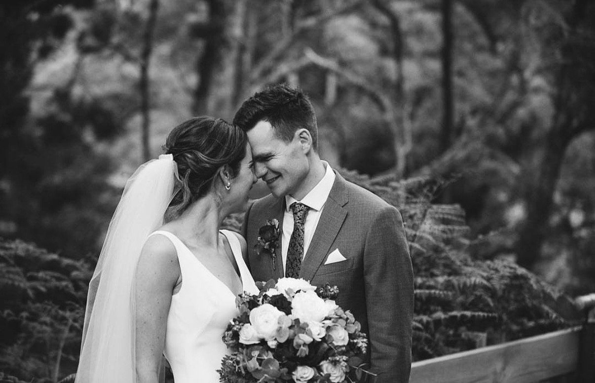 A black and white photo of a bride and groom smiling with foreheads touching, outdoors with trees in the background. The bride holds a bouquet of flowers.