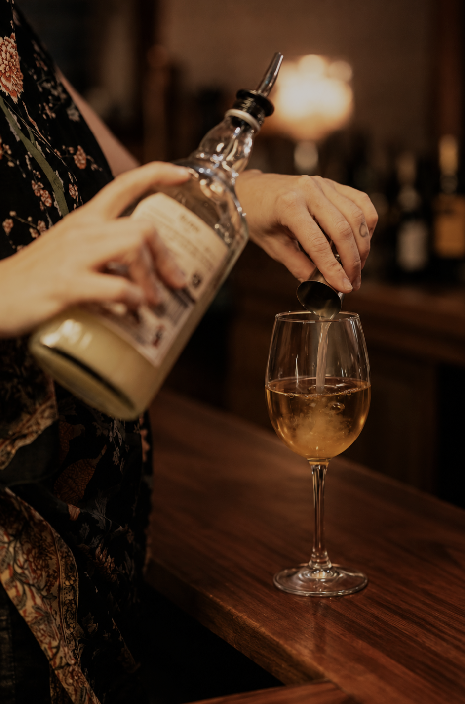 Person pouring a light-colored liquor into a wine glass on a wooden bar counter, with a blurred warm-lit background.