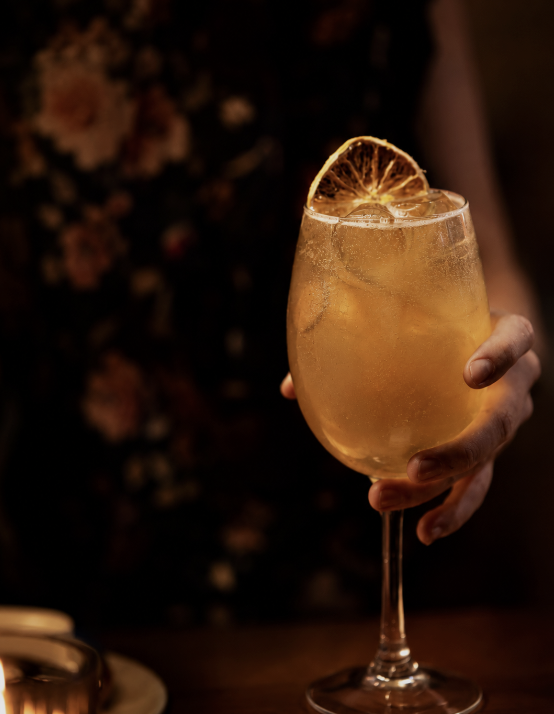 A hand holding a stemmed glass filled with a golden-colored cocktail and ice cubes, garnished with a dried lemon slice.