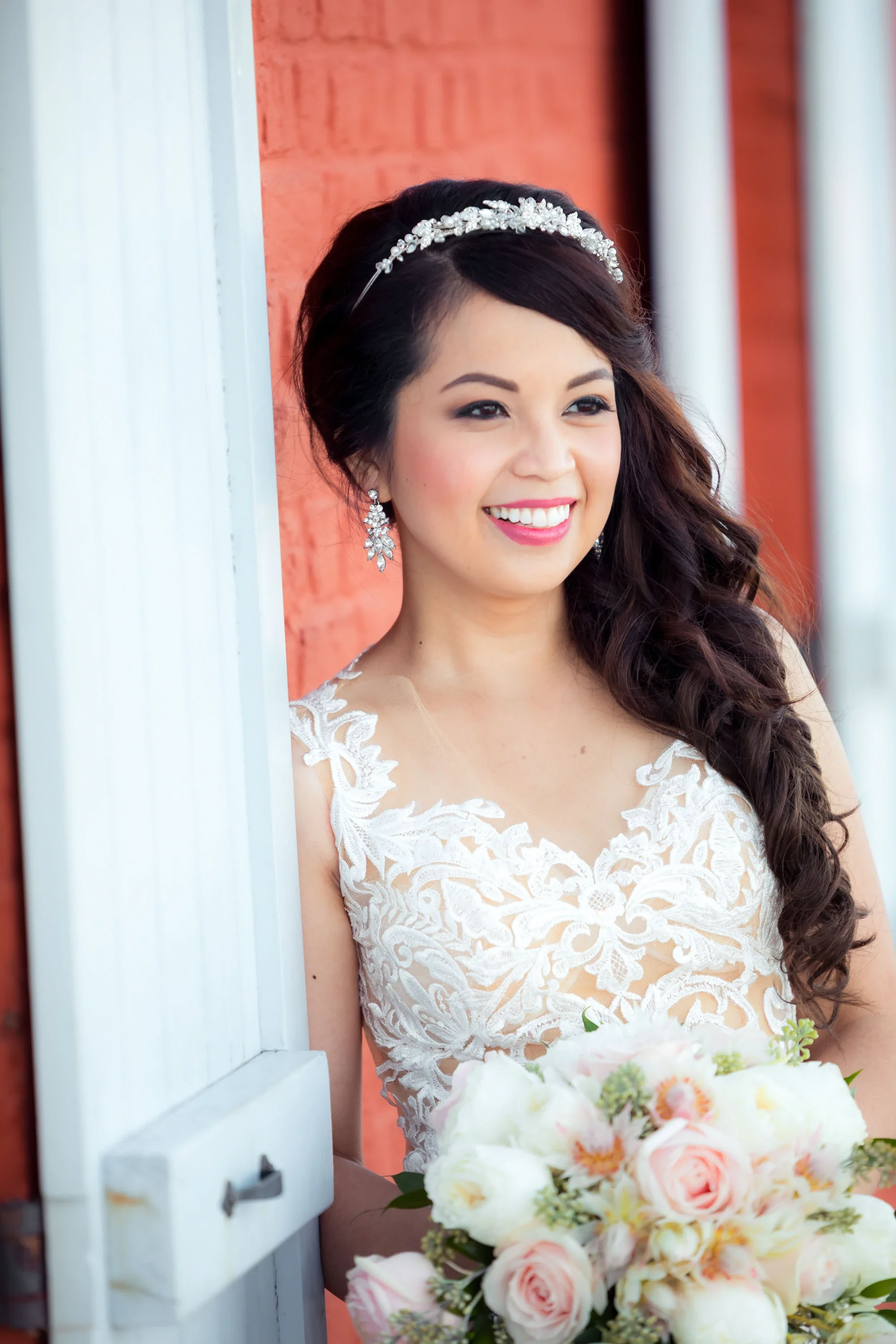 Radiant bride smiling joyfully in an intricate lace wedding gown with crystal headpiece and dangling earrings, holding a soft pink and white rose bouquet while framed by a white door against a warm red brick wall, affordable wedding photography by Sh