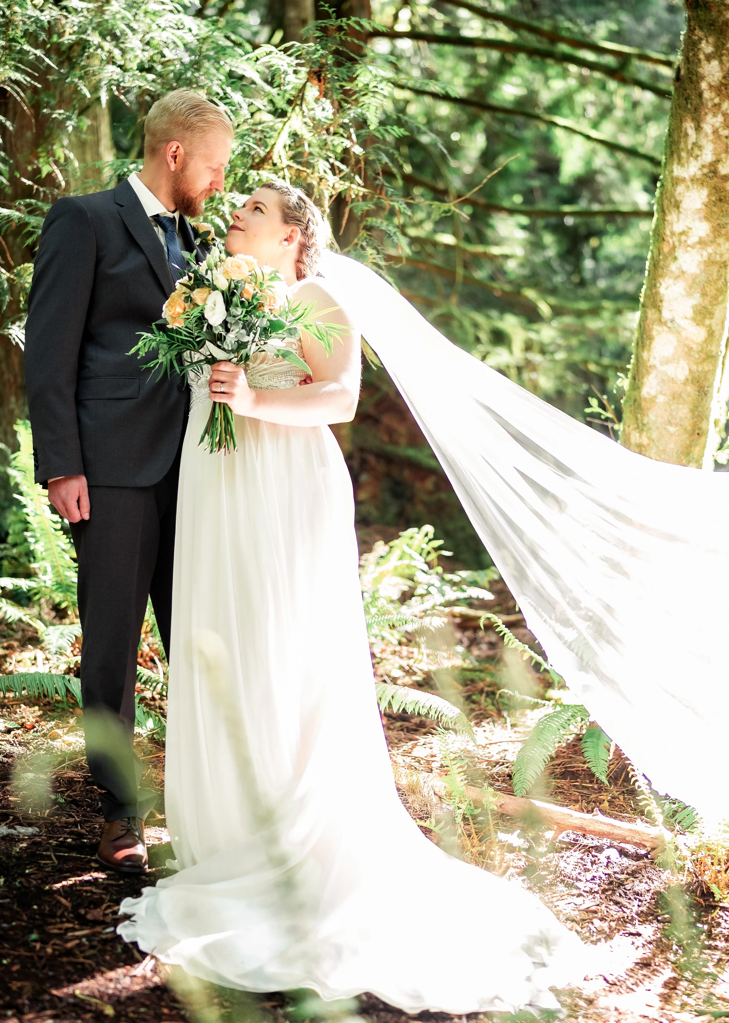 Intimate portrait of bride and groom sharing a tender moment in a lush Pacific Northwest forest, with flowing veil catching the sunlight and bride holding a natural peach and greenery bouquet, timelessly documented by an Abbotsford based wedding phot