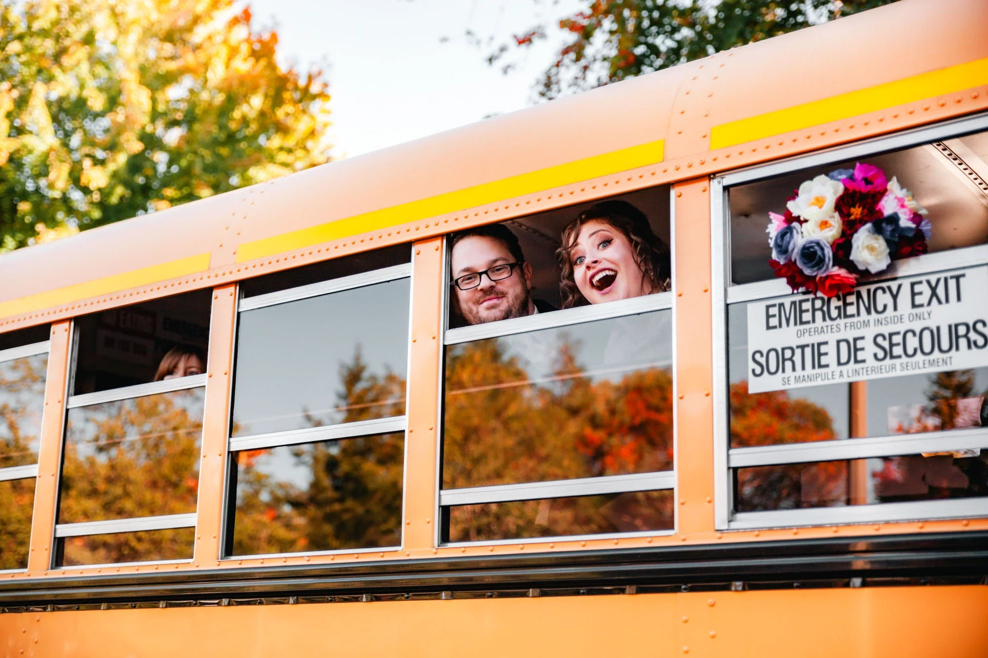 Excited bride and groom leaning out the windows of a bright orange vintage school bus, laughing joyfully with the bride waving her colorful bouquet. A whimsical and fun wedding moment captured by Shakil Hussain, Abbotsford BC wedding photographer.