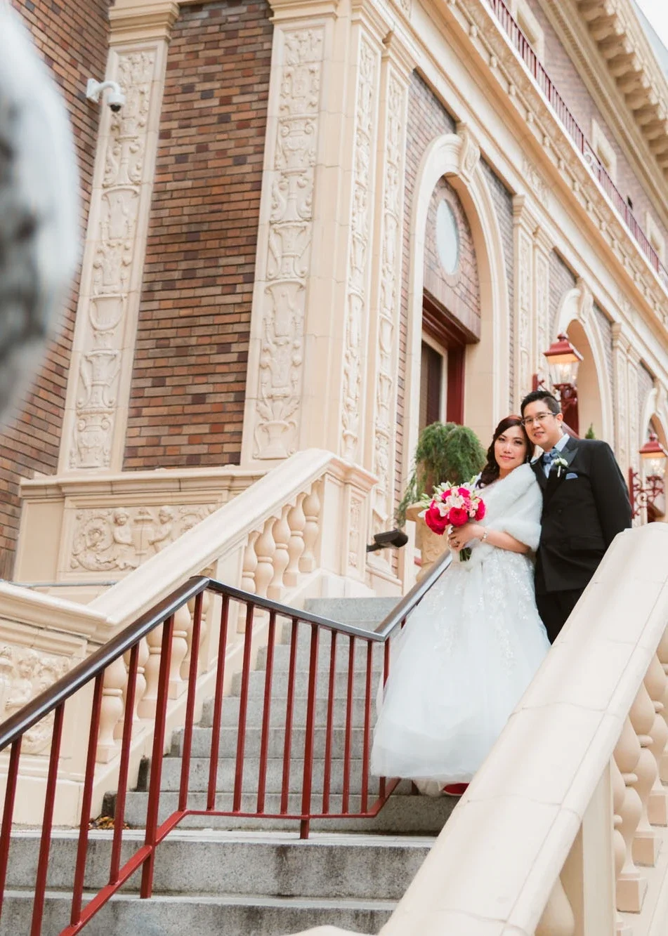Smiling bride and groom posing together on an ornate outdoor staircase with elegant architectural details and carved stone columns, bride holding a vibrant pink rose bouquet and wearing a sparkling ballgown with fur stole, captured by an Fraser Valle