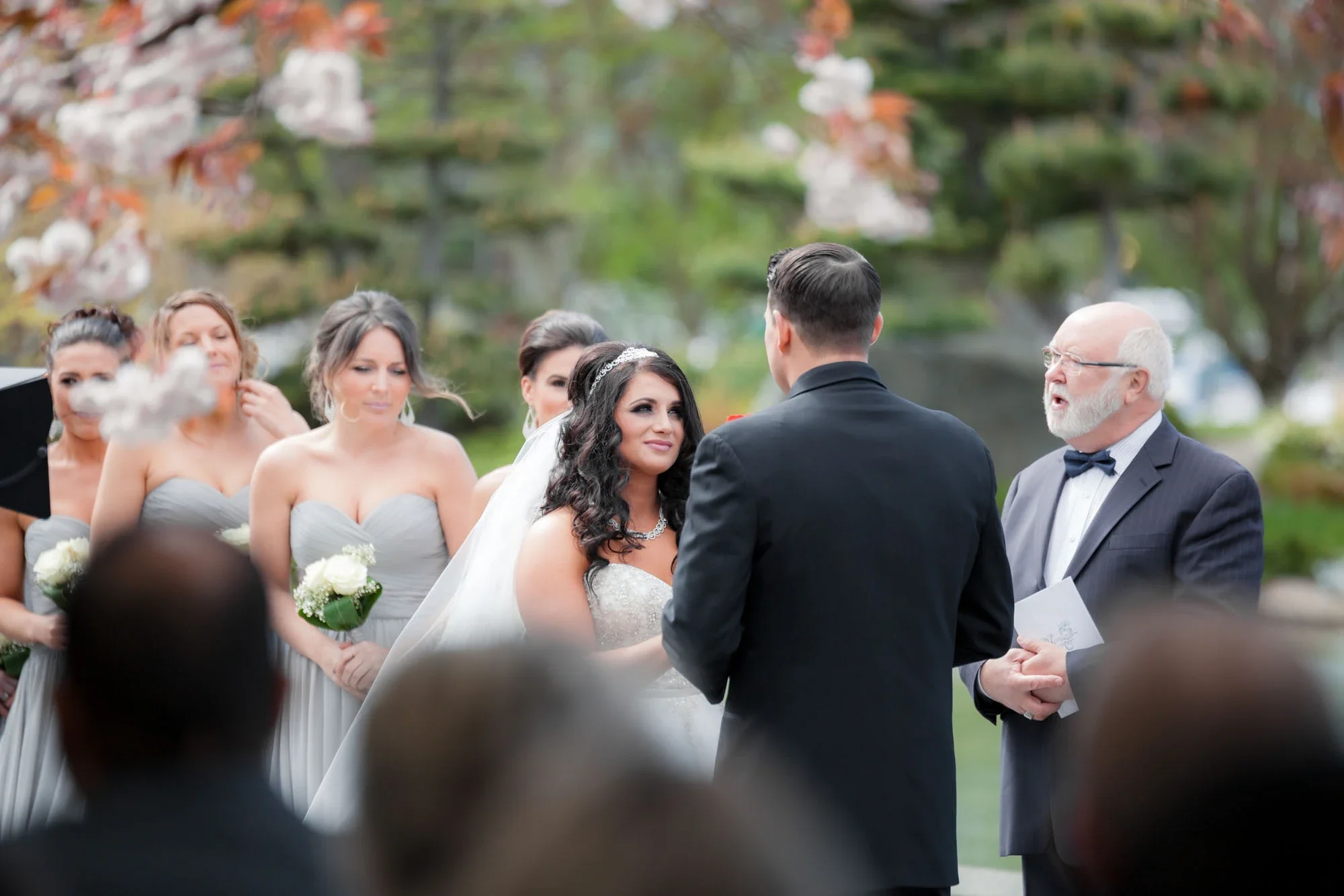 Emotional moment during an outdoor wedding ceremony in Abbotsford, BC, as the bride gazes lovingly at the groom while exchanging vows. Bridesmaids in soft gray dresses stand behind in a lush garden setting. Captured by Shakil Hussain, Abbotsford BC w