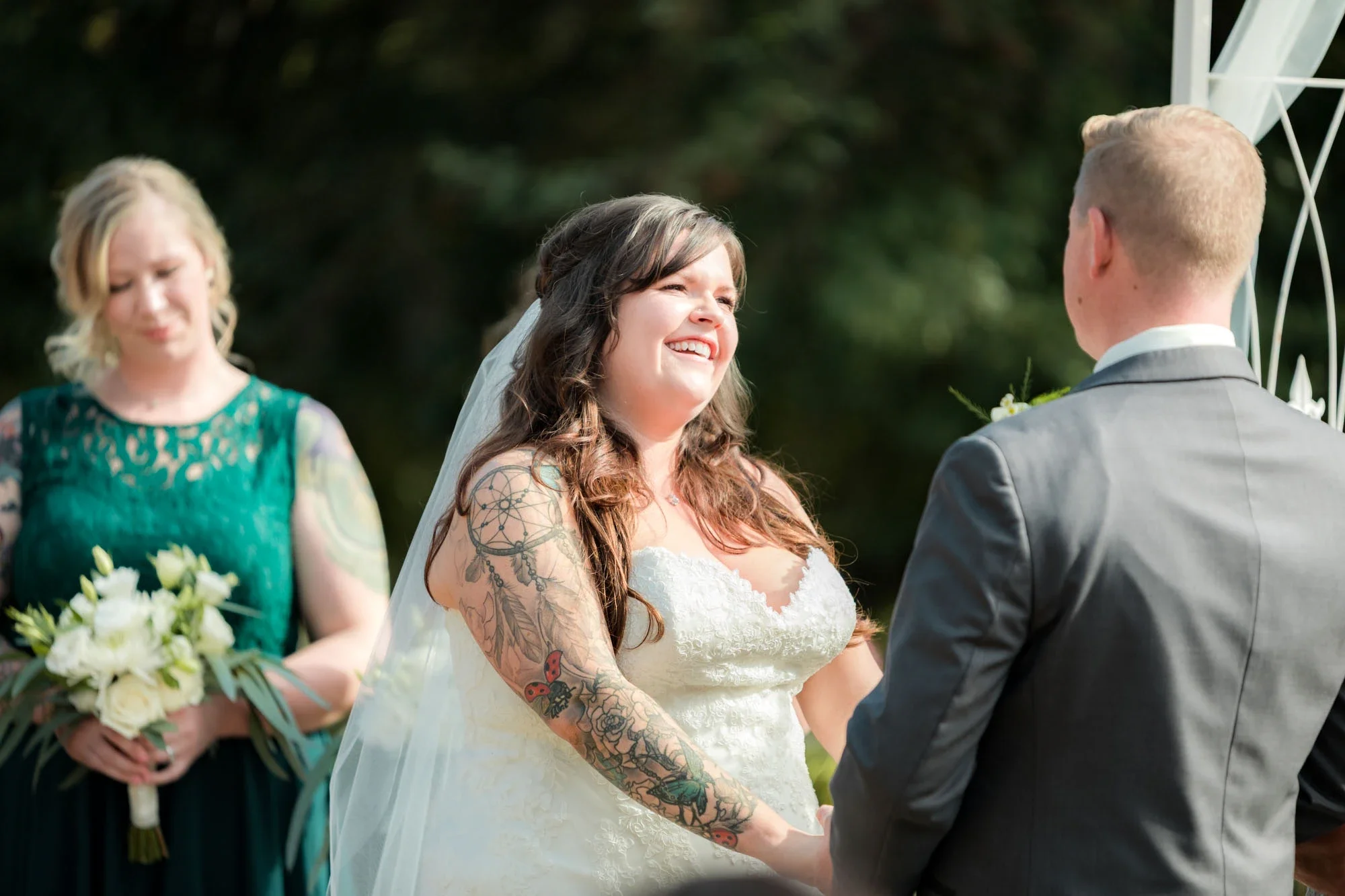 Joyful bride with visible tattoos laughing heartily during her outdoor wedding ceremony in Abbotsford BC, holding hands with the groom under a white arch. Captured by Shakil Hussain, Abbotsford BC wedding photographer.