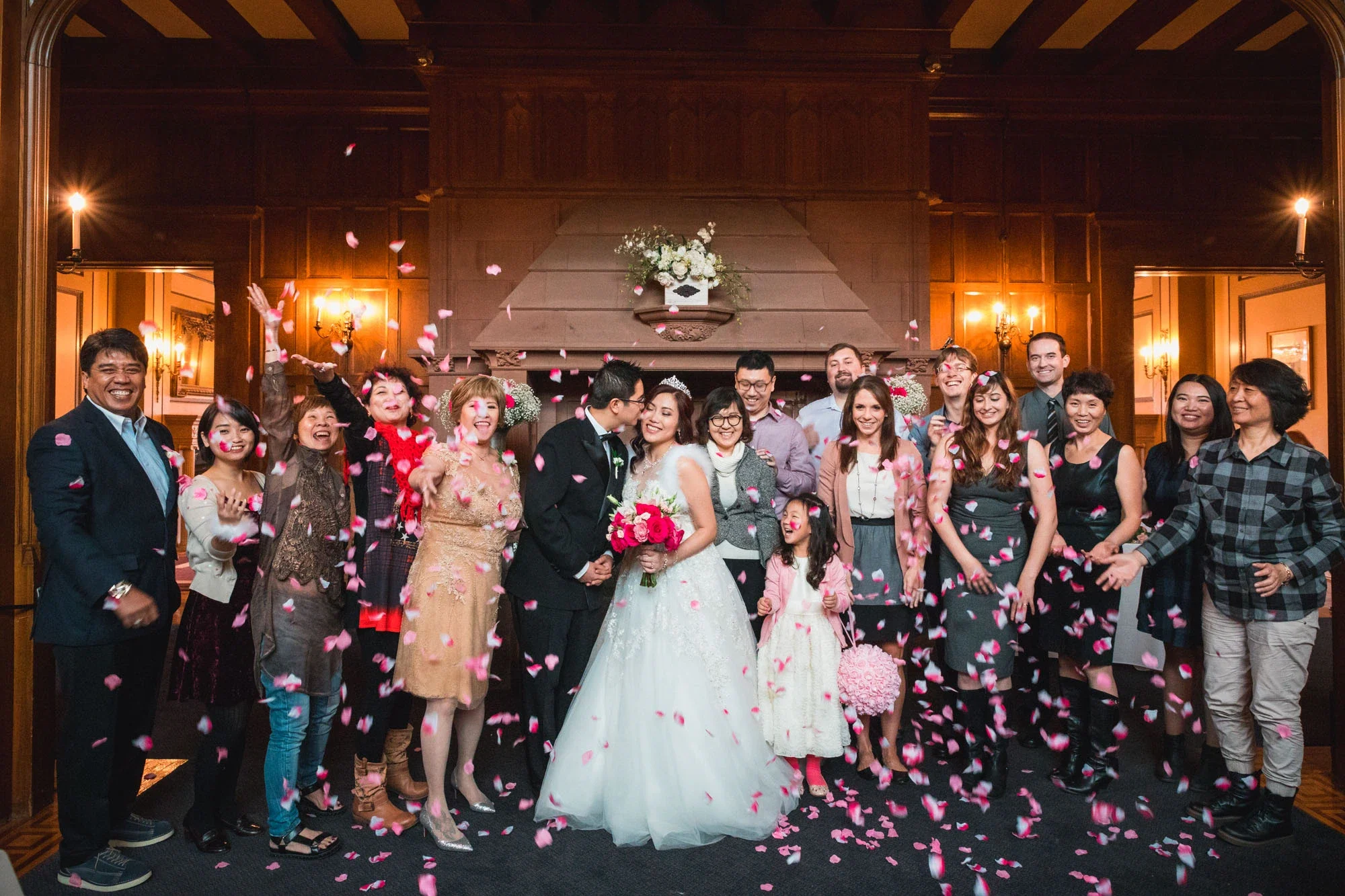 Joyful bride and groom sharing a kiss surrounded by cheering family and friends throwing pink rose petals during a festive ceremony exit in an elegant wood-paneled venue with warm lighting, affordable wedding photography by Shakil Hussain Photography