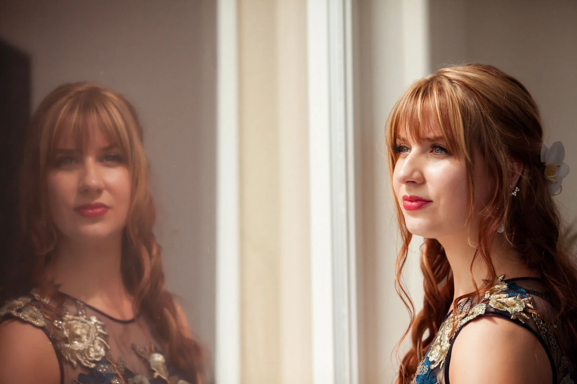 Beautiful bride with soft wavy hair and floral hairpiece gazing thoughtfully into a mirror with natural window light illuminating her intricate embroidered wedding dress during getting ready in Abbotsford BC. Captured by Shakil Hussain, Abbotsford BC