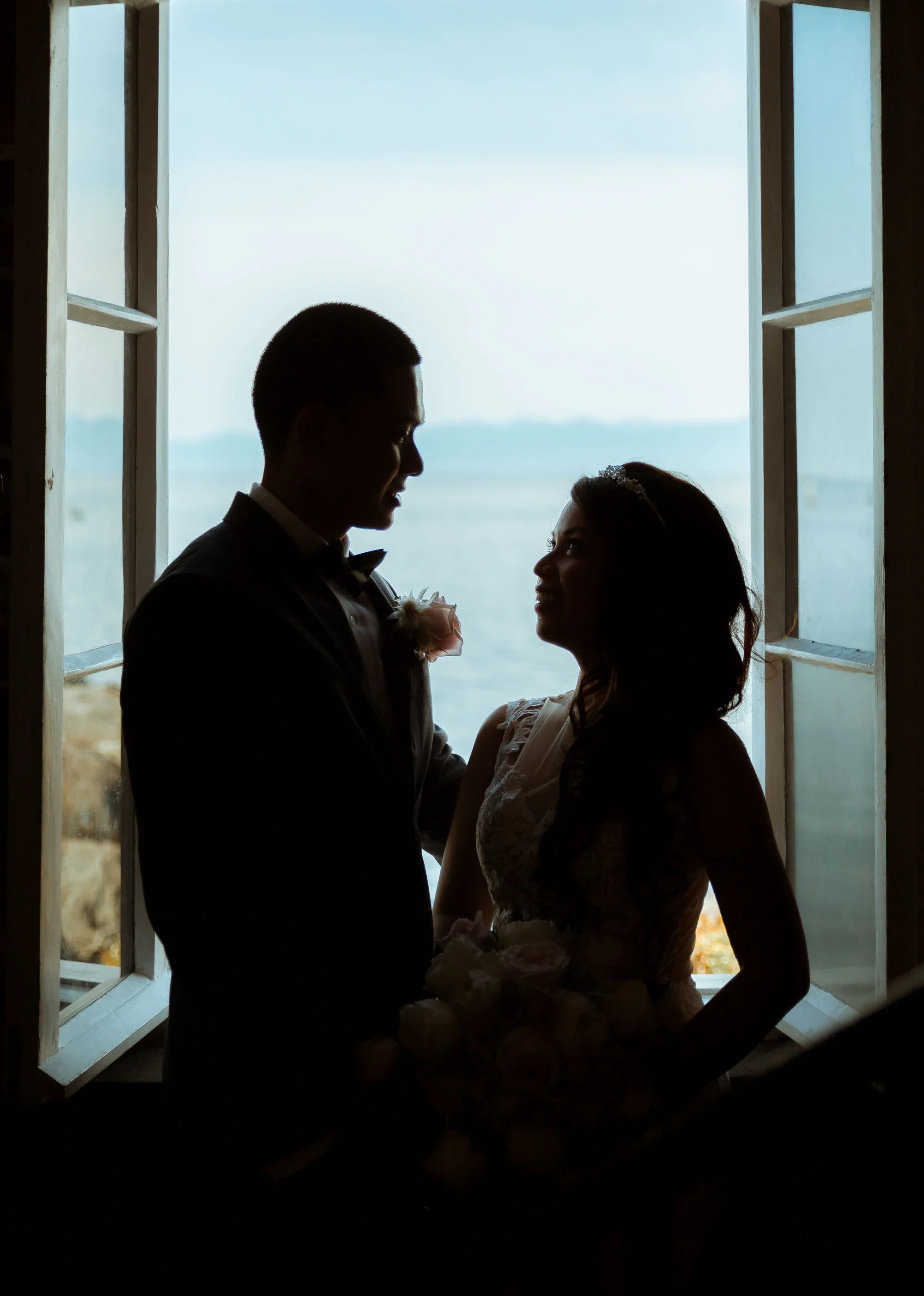 Romantic silhouette of bride Maica and groom Andrew gazing at each other in an open window overlooking the ocean, with soft natural light highlighting her lace wedding gown and bouquet, affordable wedding photography by Shakil Hussain Photography ser