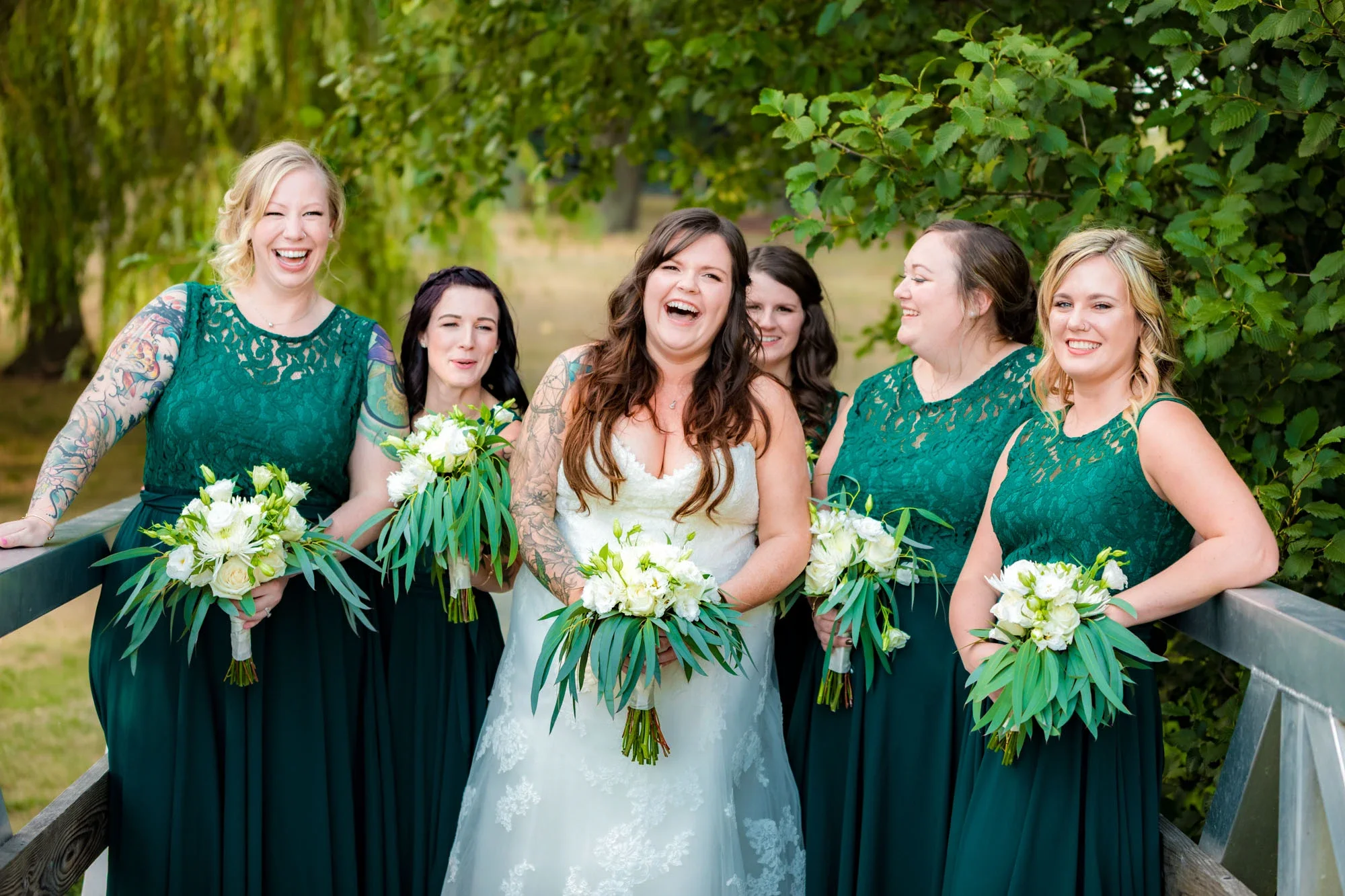 Joyful bride with visible tattoos laughing with her bridesmaids in matching emerald green lace dresses, holding white and greenery bouquets outdoors under lush trees in Abbotsford BC. Captured by Shakil Hussain, Abbotsford BC wedding photographer.
