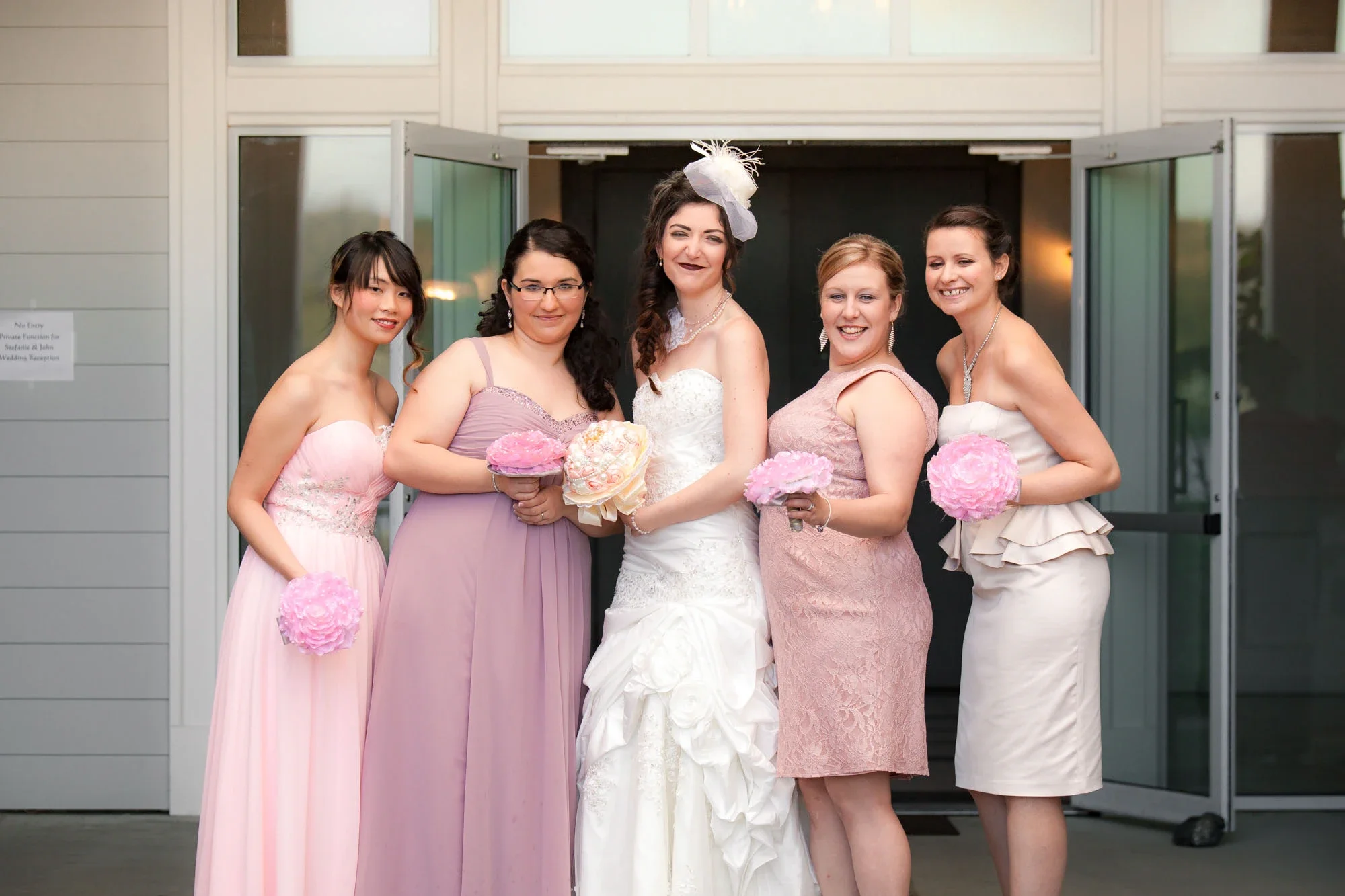 Smiling bride in a white ruffled wedding gown surrounded by her four bridesmaids in coordinating pastel pink and mauve dresses, each holding pink floral bouquets, posing together outside the venue in Abbotsford, British Columbia.