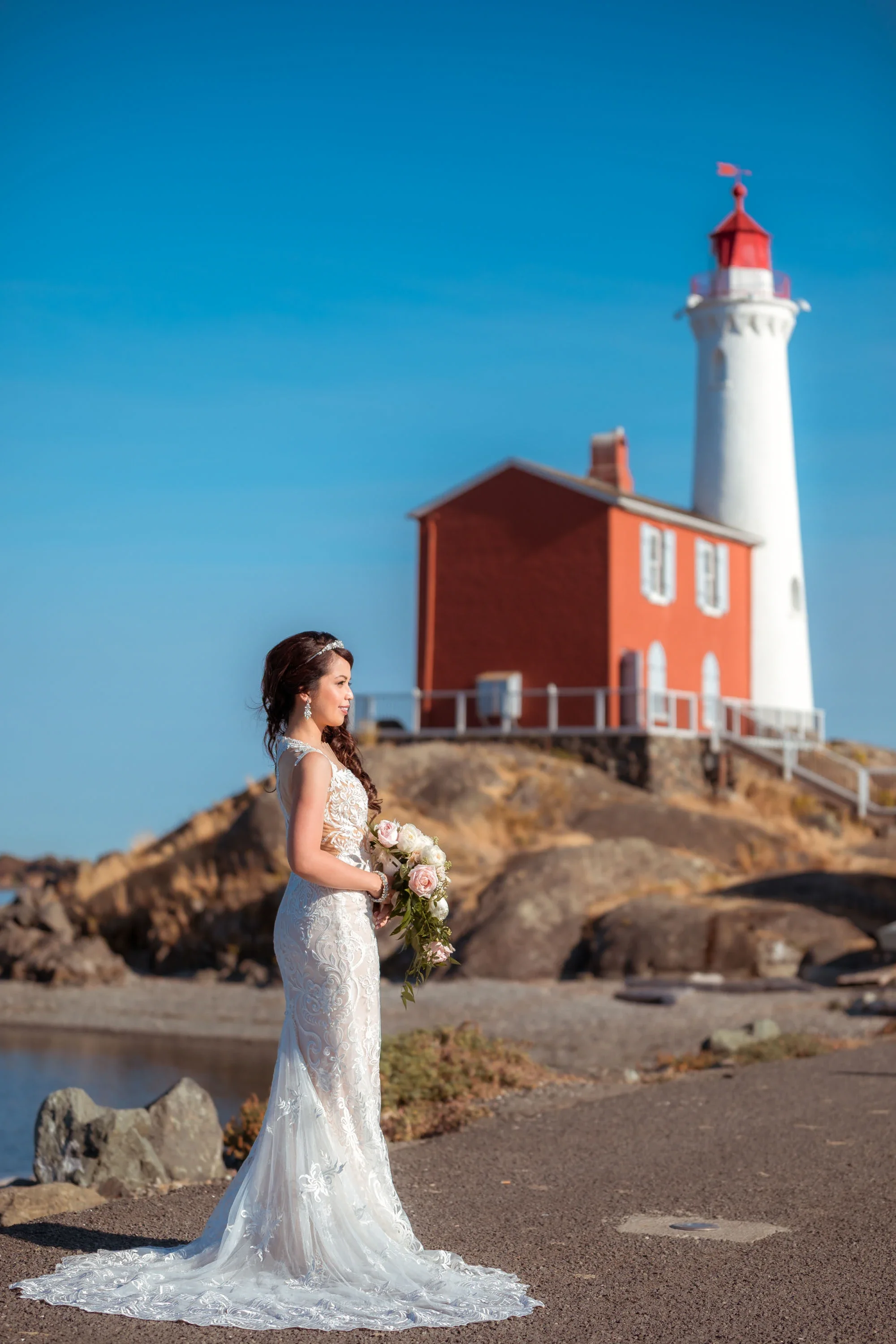 Elegant bride in a fitted lace wedding gown with long train and crystal tiara holding a soft pink and white rose bouquet, gazing thoughtfully toward the ocean with a historic red and white lighthouse in the background under clear blue skies, affordab